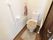 部屋のトイレである。奥の壁にはL字型の手すりが付いている。そばには緊急呼び出しボタンや温水便座洗浄の操作パネルがある。