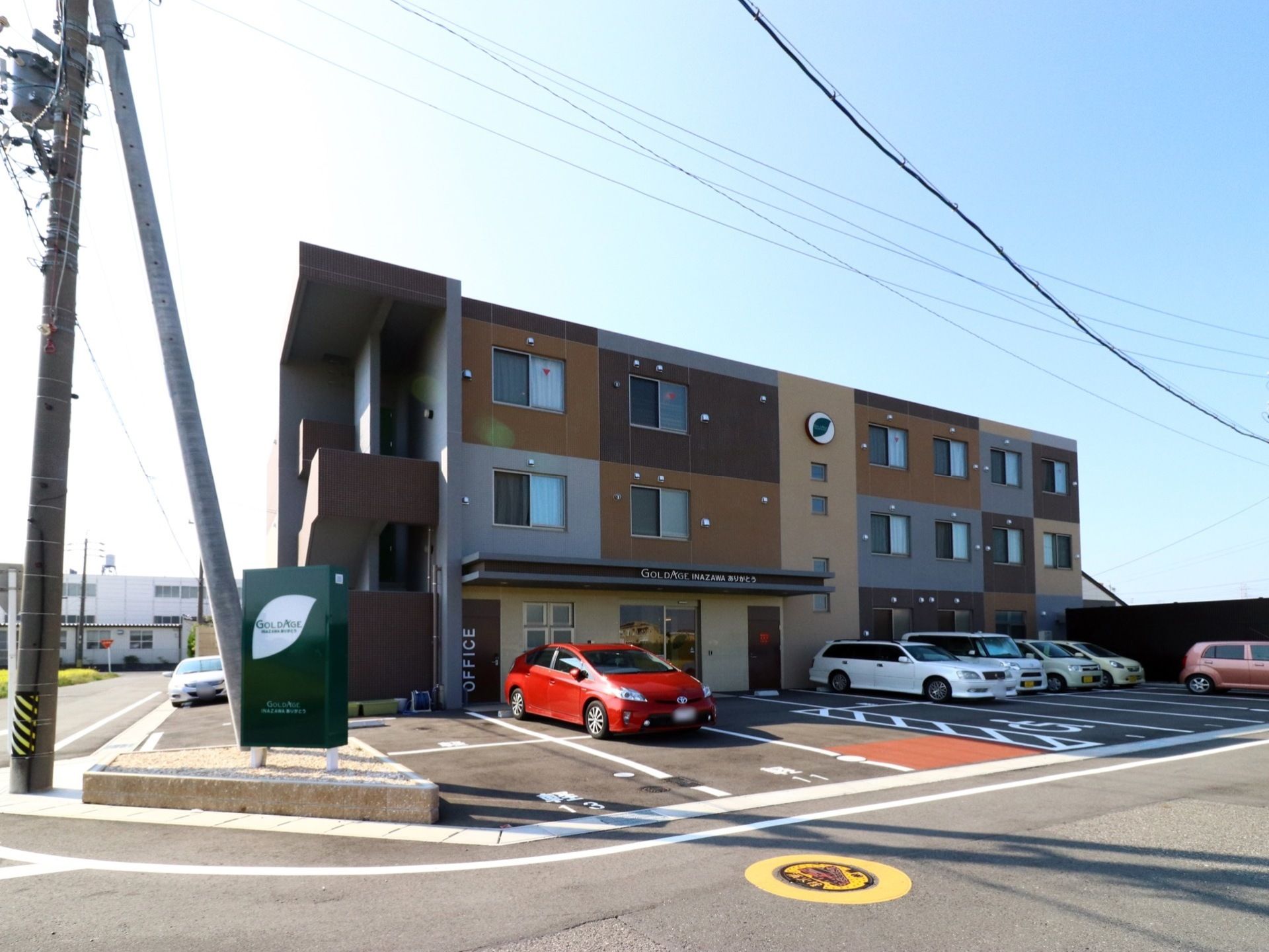 3階建ての施設で手前に看板が立っている。非常用階段が左側に設けられている。広い駐車場もあるので多くの車が収容できる。