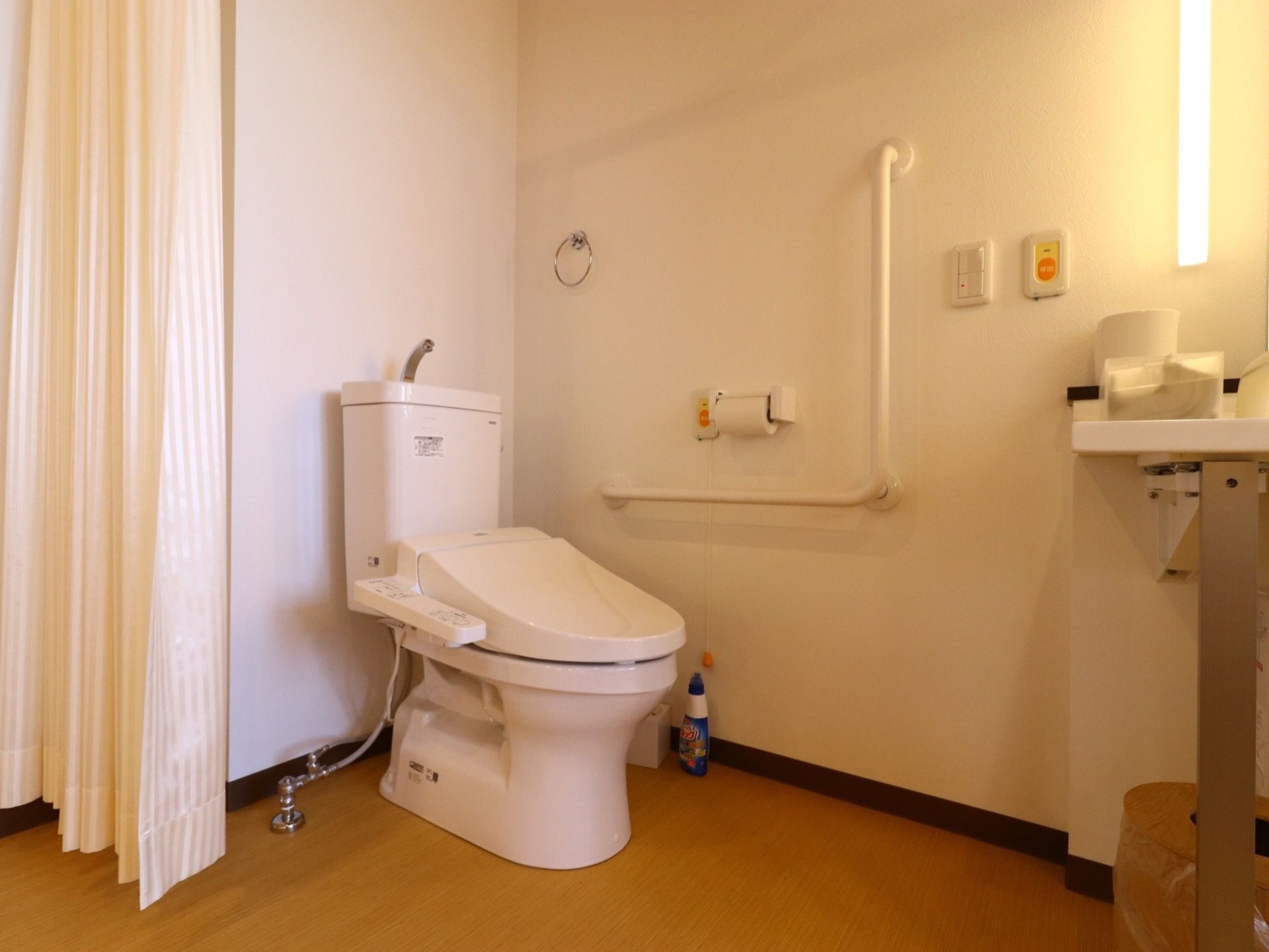 車椅子仕様のトイレである。L字型の手すり、呼び出しボタンが壁に取り付けられている。カーテンで仕切られている。