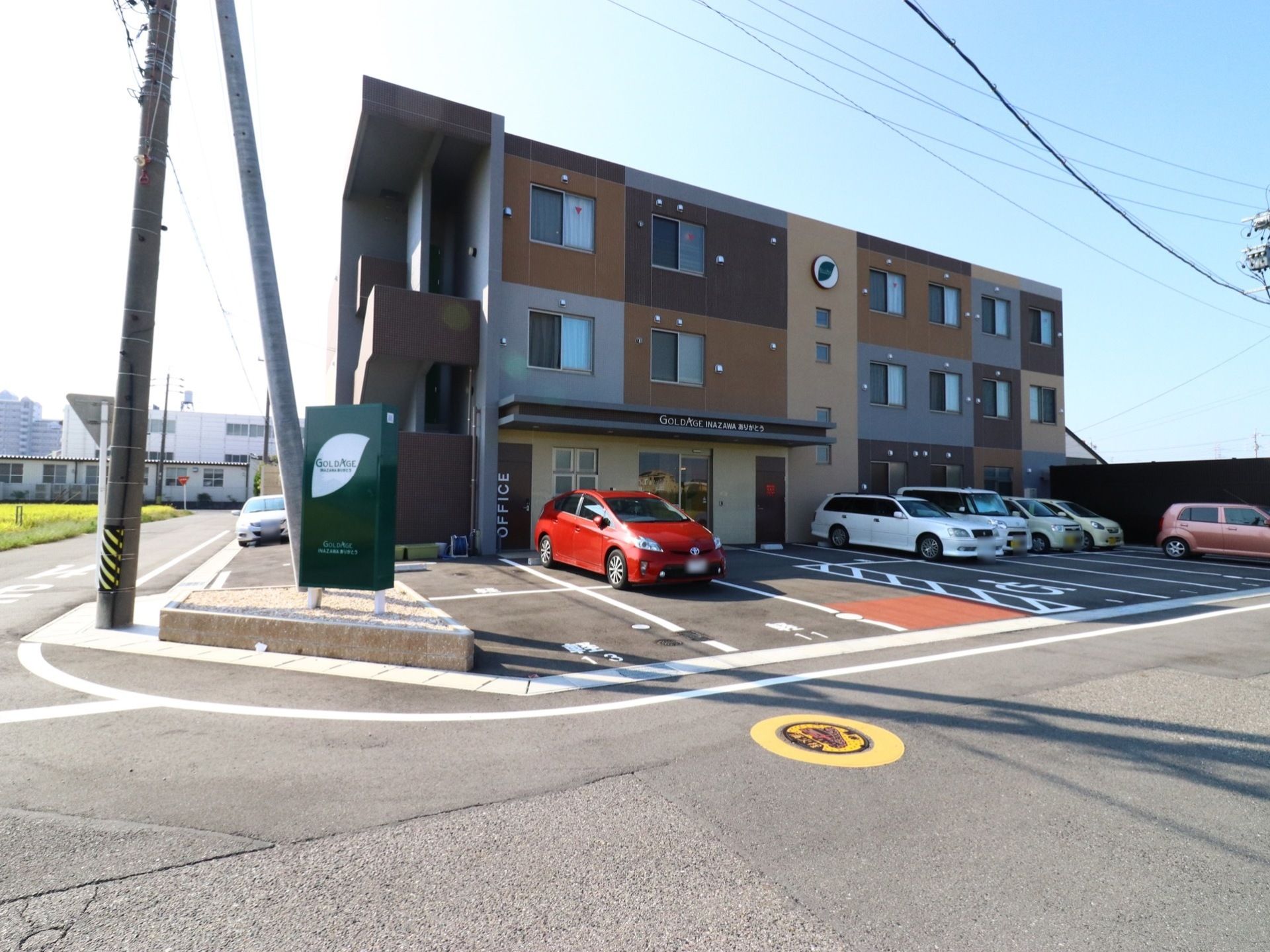 施設外観は、ブラウン基調の3階建ての建物になっていて、緑色の看板が立っている。駐車場も設けられている。