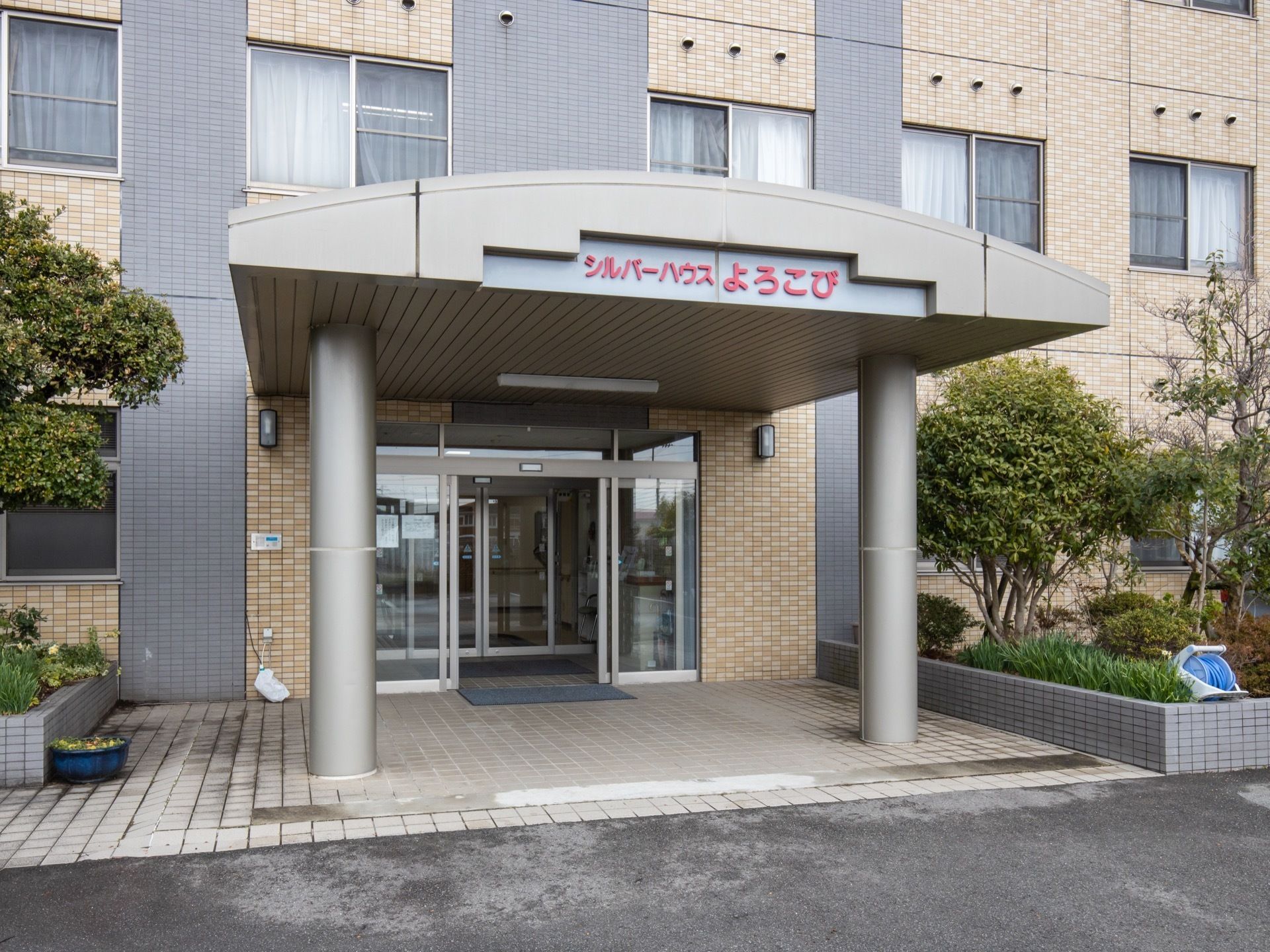 玄関の扉はガラス張りの自動ドアで、床はフラットなタイル敷きである。上部には大型の屋根を設置し、前面を太い柱で支えている。