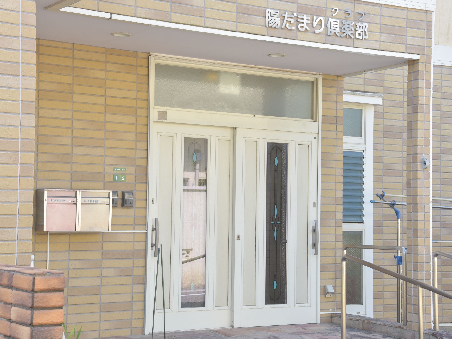施設の建物は1階部分の外壁はタイル張りになっており、通路には手摺りが設置されている。玄関ドアの横にはインターフォンが2つと郵便受けが2つ。