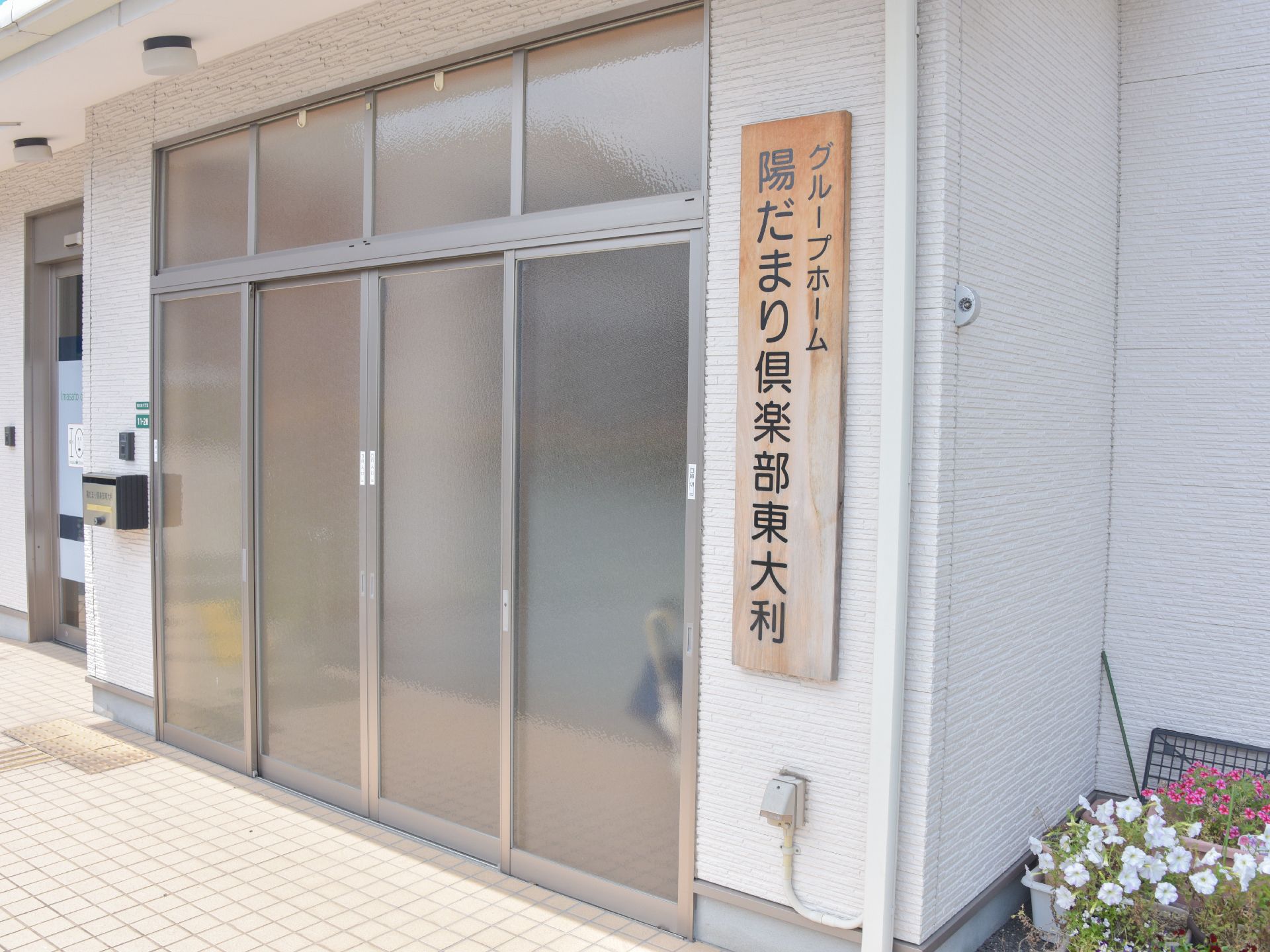 曇りガラスのドアの並んだ広々とした施設の入り口。右側には木の板に黒い文字で施設の名前が書かれた表札がある。