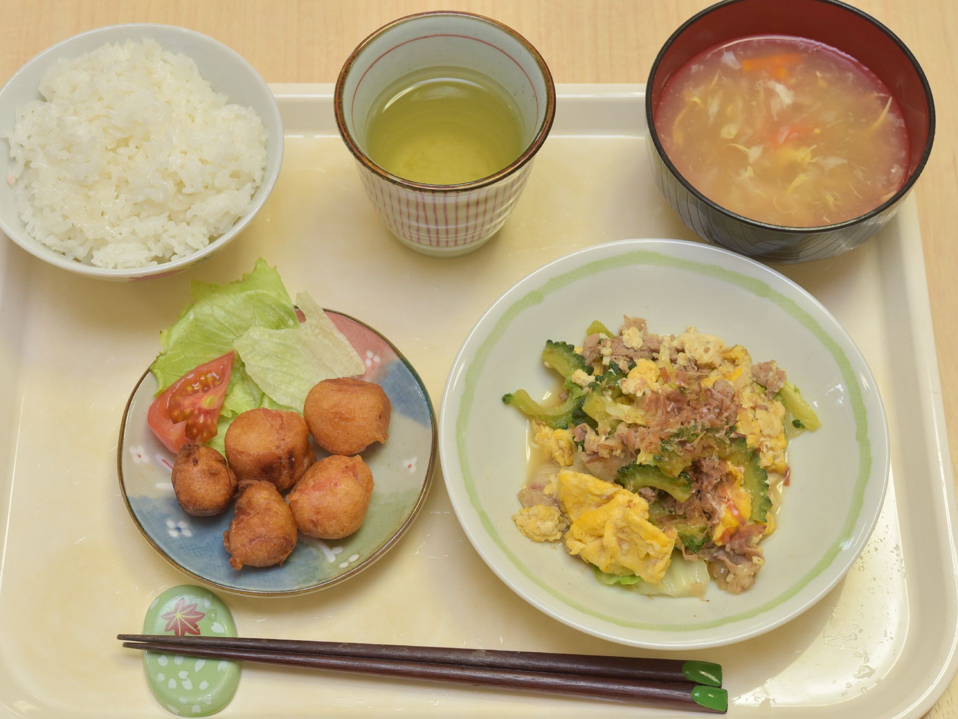お盆の上に並べられた施設の食事。ご飯とお汁のお椀におかずが2皿ついていて、お箸とお茶が添えられている。彩にも気を配ったメニューになっている。
