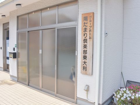 曇りガラスのドアの並んだ広々とした施設の入り口。右側には木の板に黒い文字で施設の名前が書かれた表札がある。
