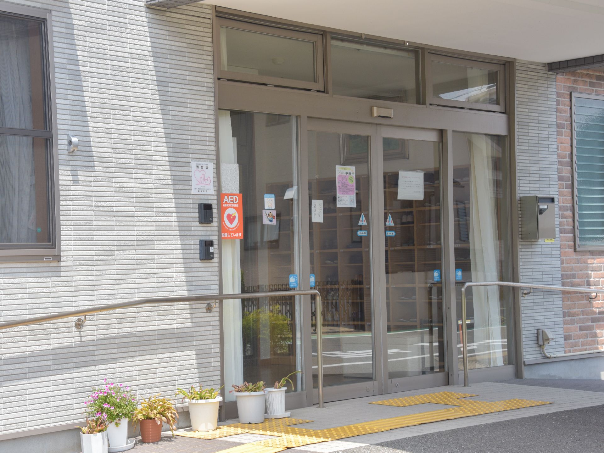 玄関のドアの両側に手すりが設けられている。また地面には点字ブロックが設置されている。鉢植えの花が飾られている。