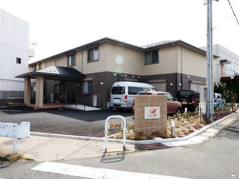 施設は白と黒の壁の二階建ての広めの一軒家というイメージの佇まい。敷地には駐車場があり塀には施設名とロゴが入ったパネルがある。