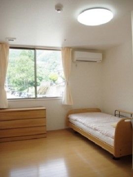 白い壁に茶色のフローリングの居室である。大きな窓なので明るい。窓からは木々を眺めることができる。ベッドと棚、エアコンが設置してある。