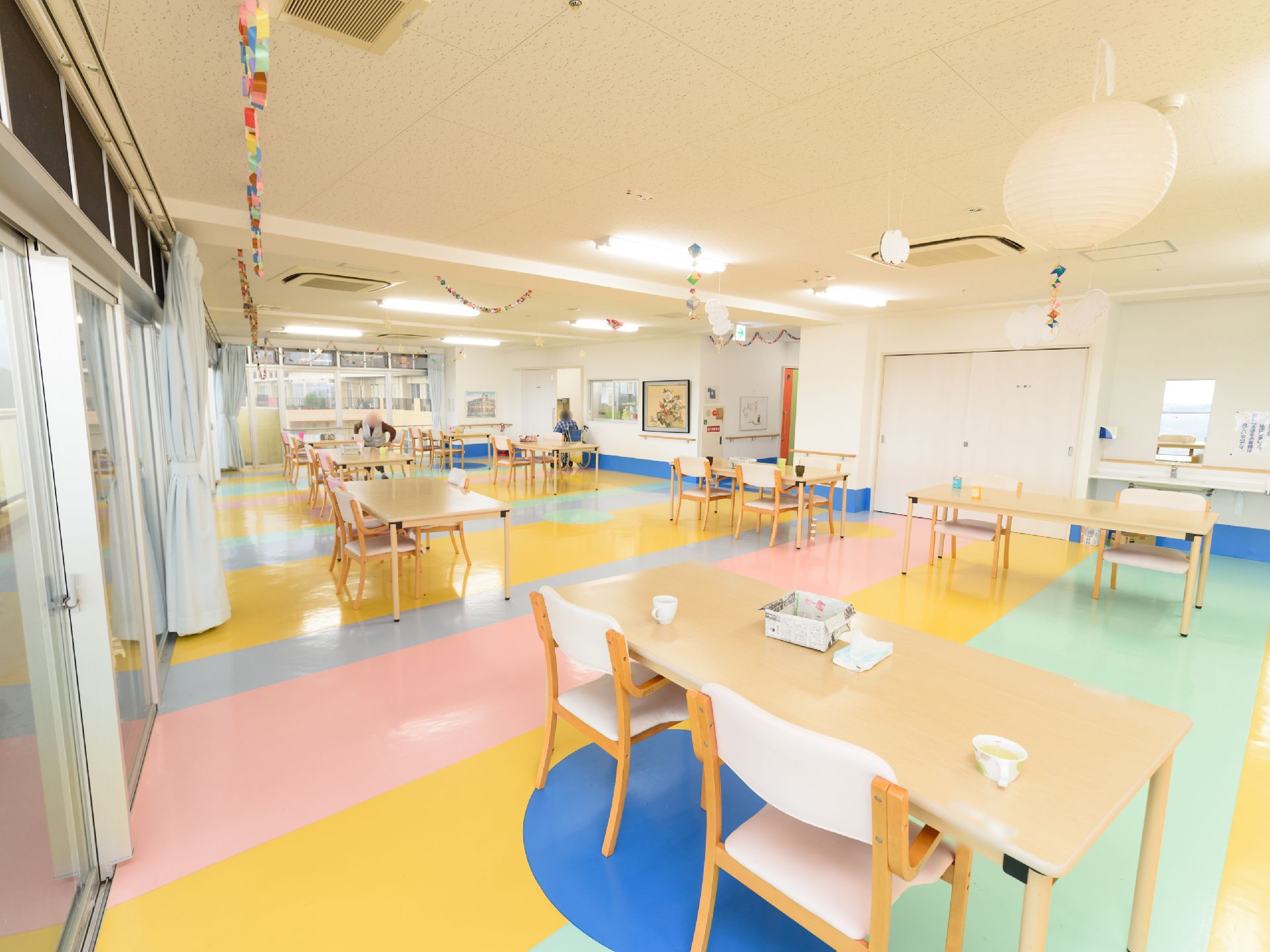 共有スペースは、明るくカラフルな床面になっている。横長のテーブルと椅子が広い間隔でたくさん置かれている。