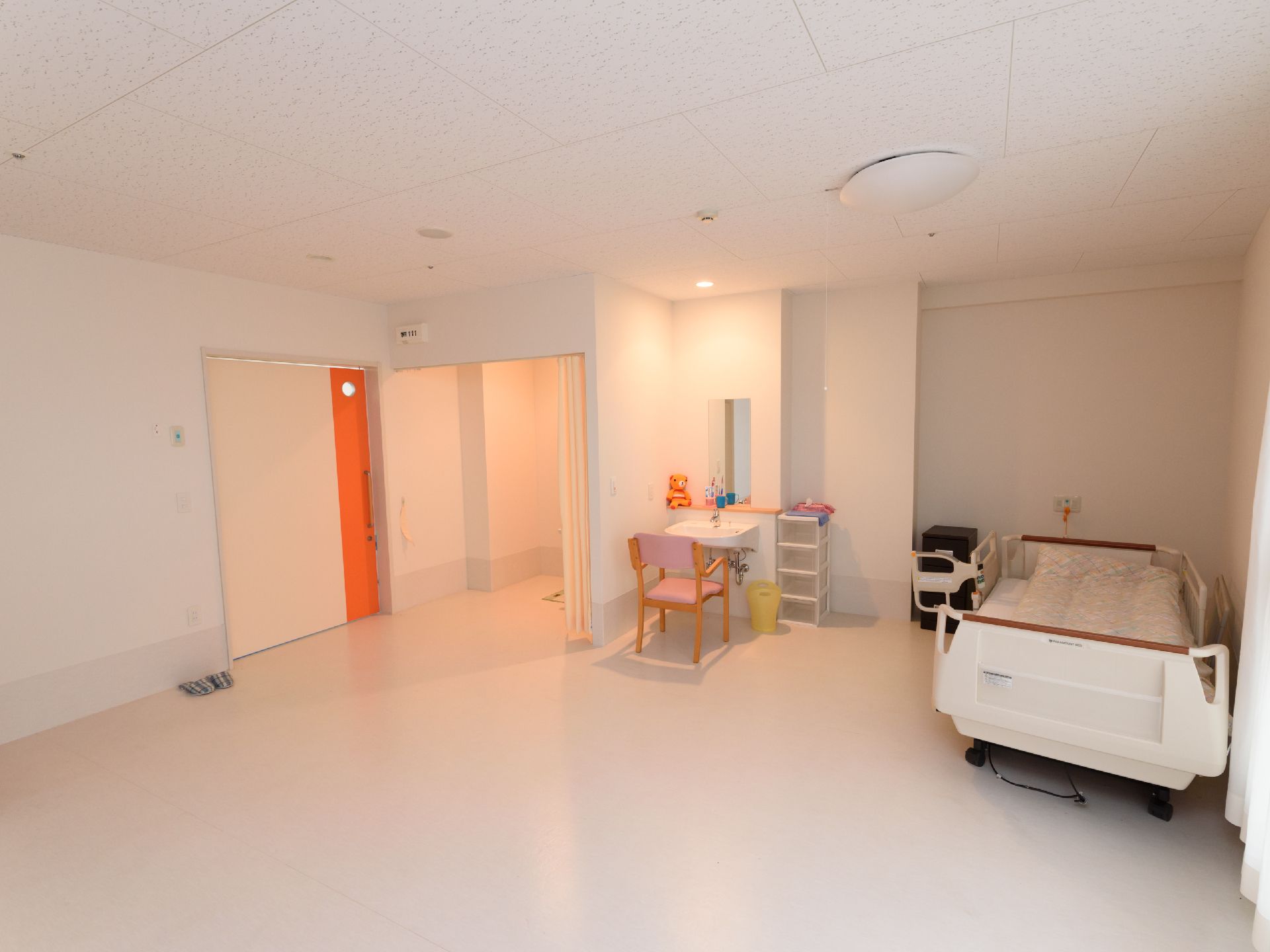 床面や壁、天井ともに白色を基調とした、とても広い居室である。介護ベッドや洗面台、収納ラックが設置されている。