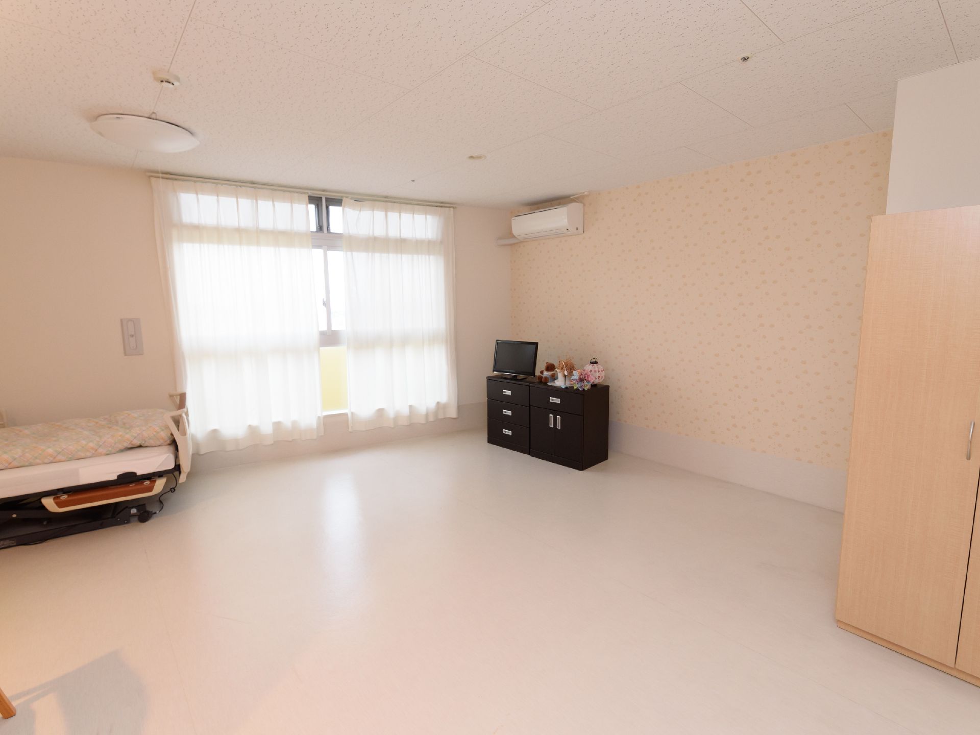 白色を基調した、とても広い居室になっている。背の高い窓の横に、介護ベッドやエアコン、テレビが設置されている。