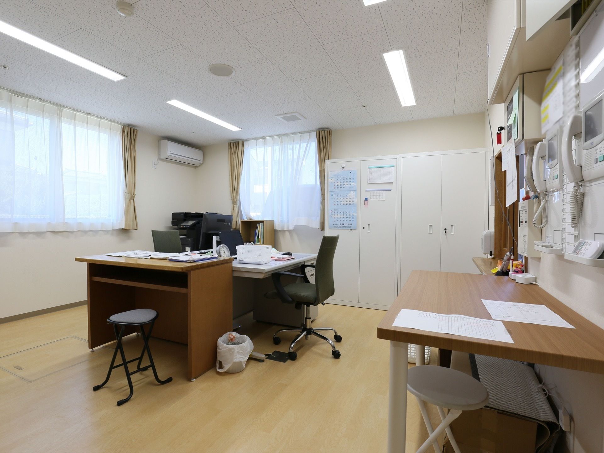 室内に、事務机や椅子が複数置かれている。奥の壁際に収納庫があり、手前の壁にインターホンが備わっている。