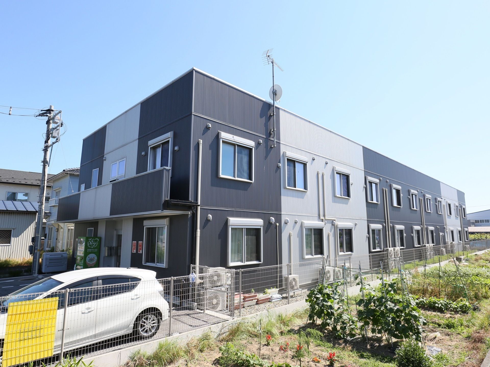 ２階建ての横に長い建物で、外壁が白と黒のツートンカラーである。玄関前に、駐車スペースや自動販売機がある。