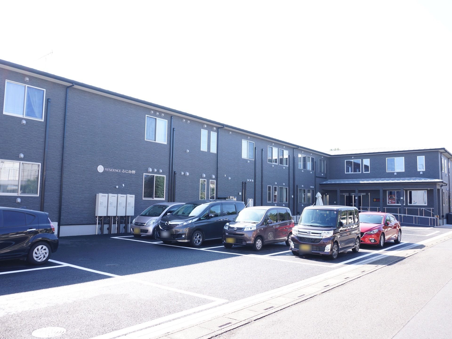 2階建ての施設外観は黒色である。敷地内には広い車の駐車スペースがあり、6台ほどの車が止められている。