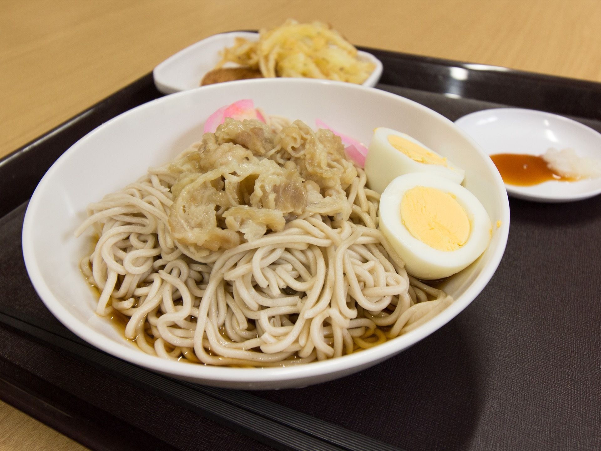 冷やし肉蕎麦がもられた食事である。天ぷら・醤油も盛りつけられている。蕎麦には茹で卵やピンクのかまぼこものっている。