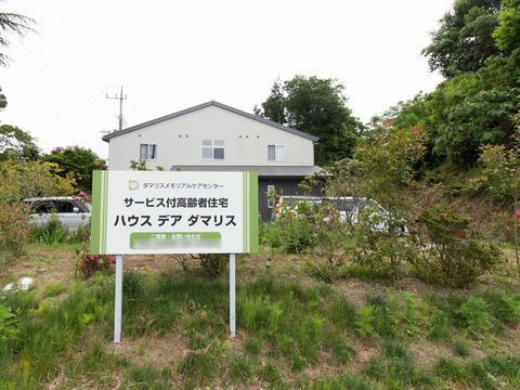 施設の前方に花壇があり、その真ん中あたりに横長の看板が立っている。草木の奥のスペースには、数台の車が置いてある。