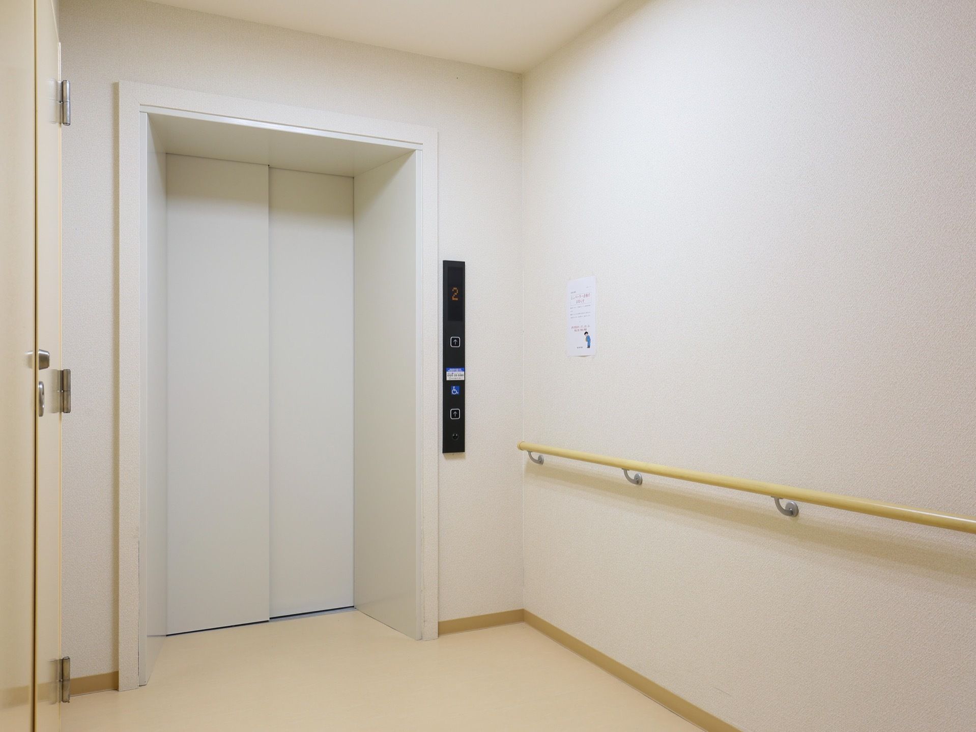 エレベーターが1台設置されており、エレベーターの右側に操作パネルがある。右側の白い壁に張り紙が貼られている。