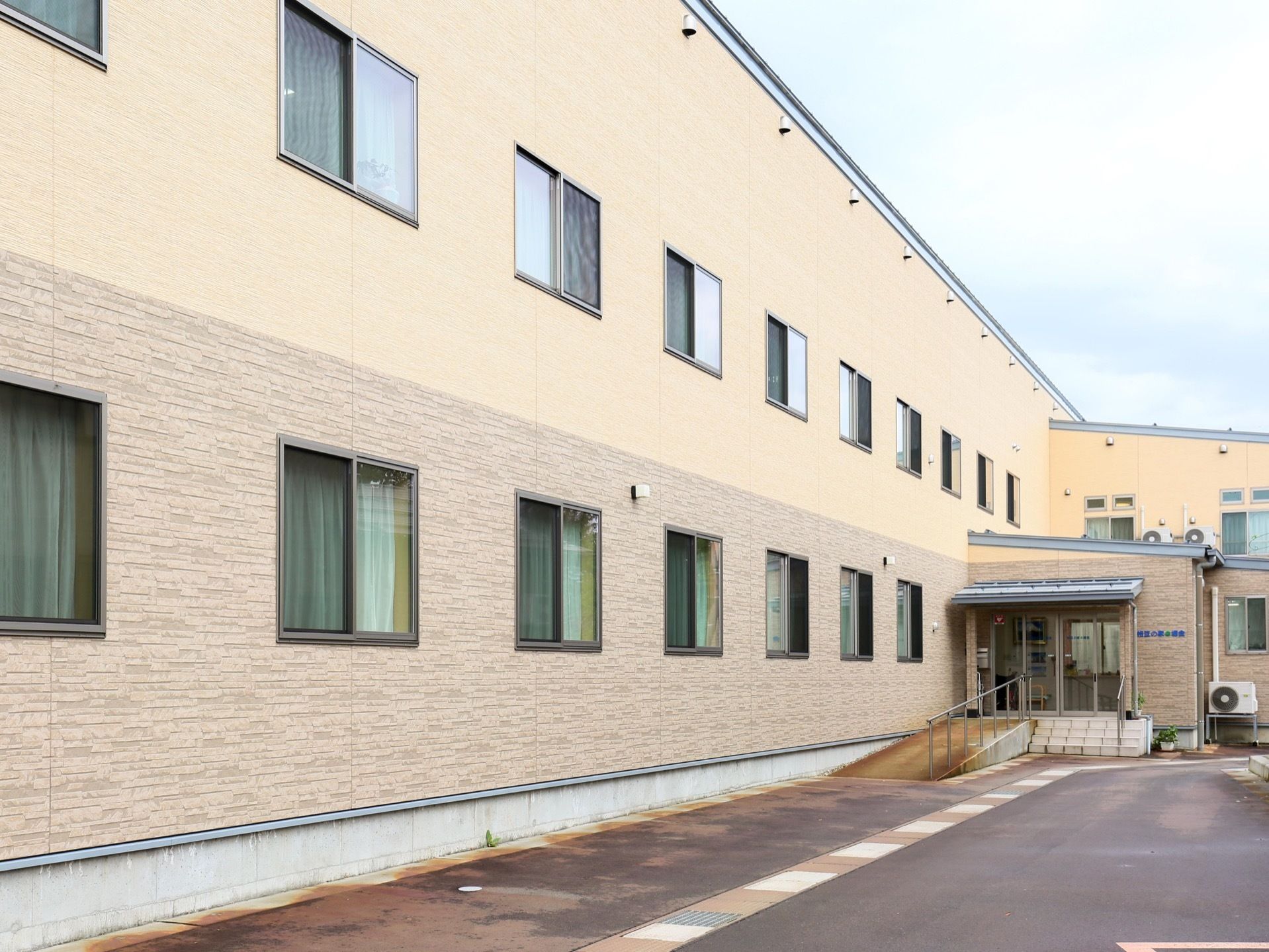 1階と2階で異なる色が使用された外壁の建物で、玄関前には階段とスロープが設置されている。階段の右側に鉢に入った植物が置かれている。