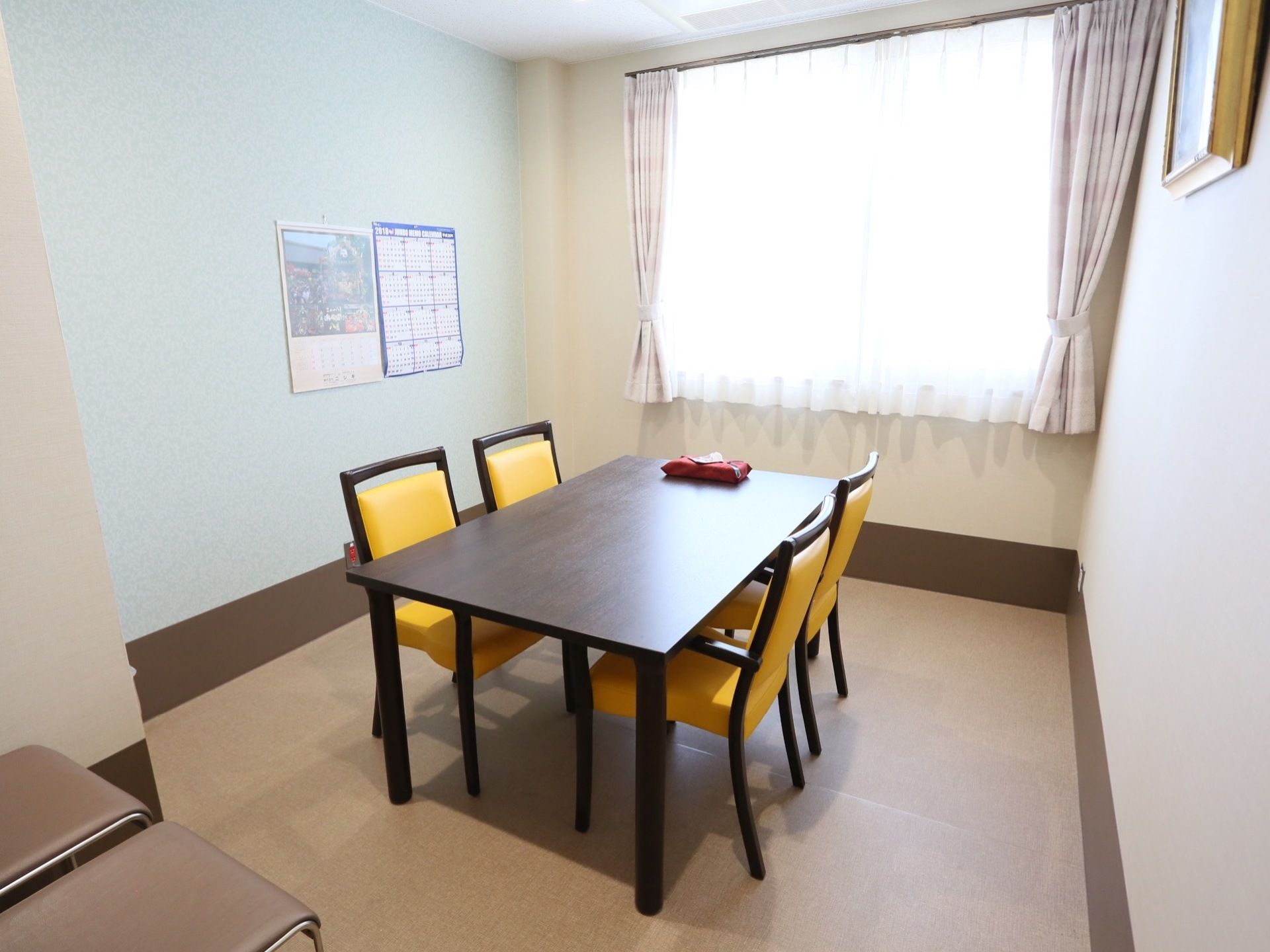腰窓のある明るい部屋の中央に、木製テーブルと４脚の椅子が置かれている。壁にはカレンダーと絵画がかかっている。