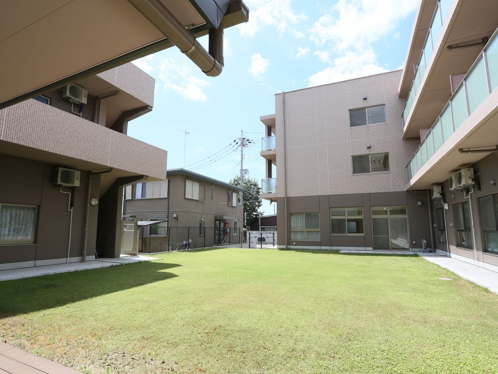 青い芝生の中庭を囲むように施設が建っている。３階建ての建物には半透明のパネル式のフェンスが設けられ、エアコンの室外機が設置されている。
