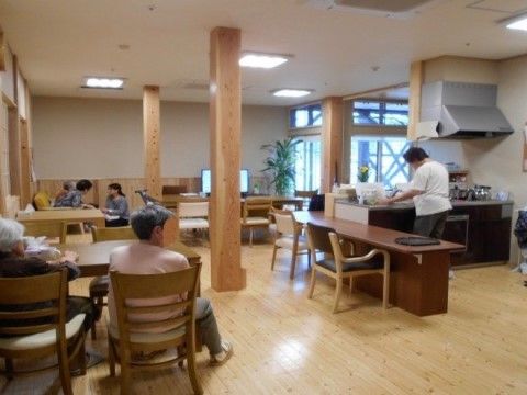 木のぬくもりを活かしたゆったりとした空間にテーブルと椅子が並べられている。オープンタイプのキッチンが設置されている。