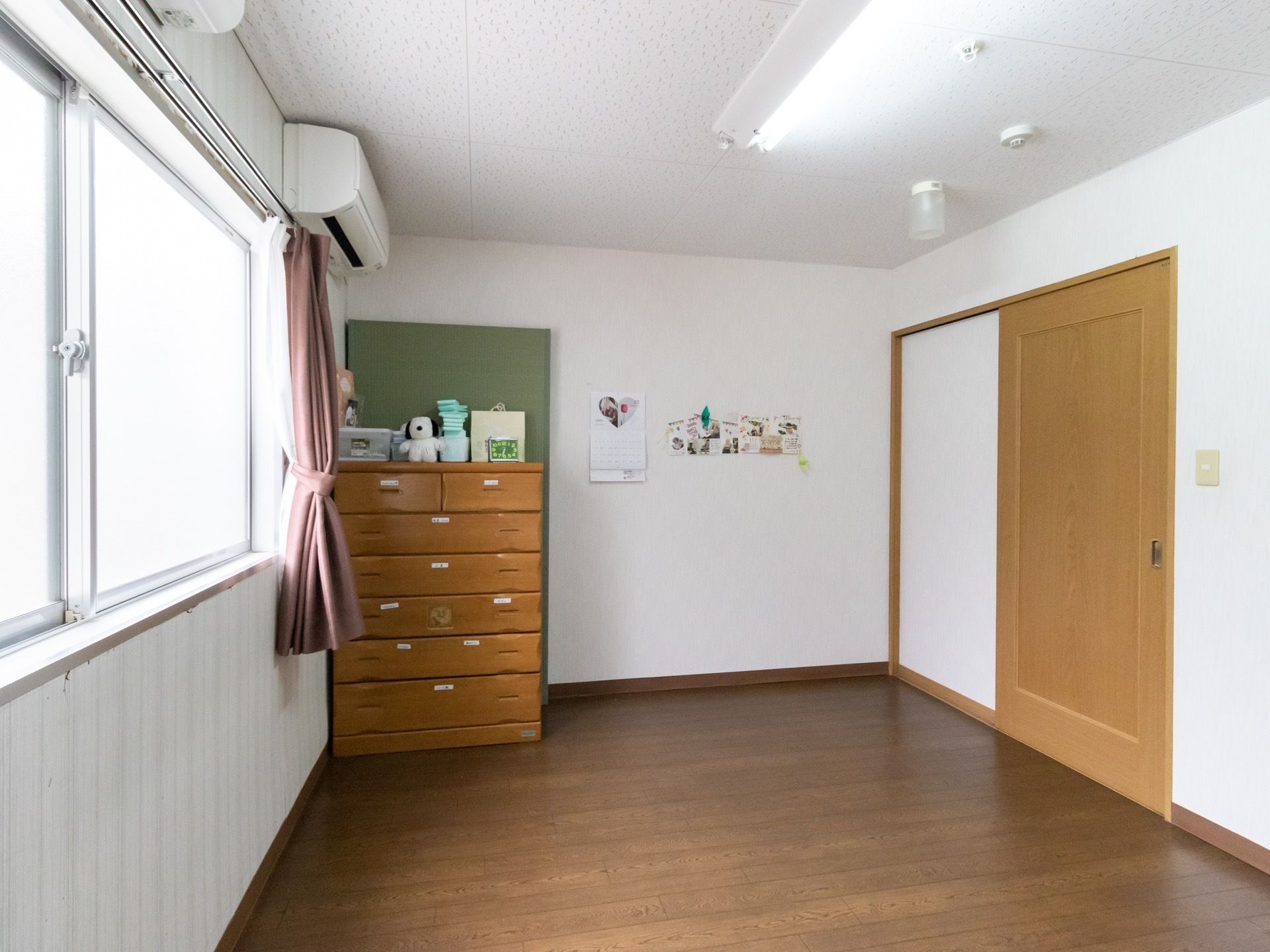 窓のすぐ側には、木製のタンスが置いてありその上には小物が飾られている。窓には、白いレースカーテンとともに茶色のカーテンが付けられている。
