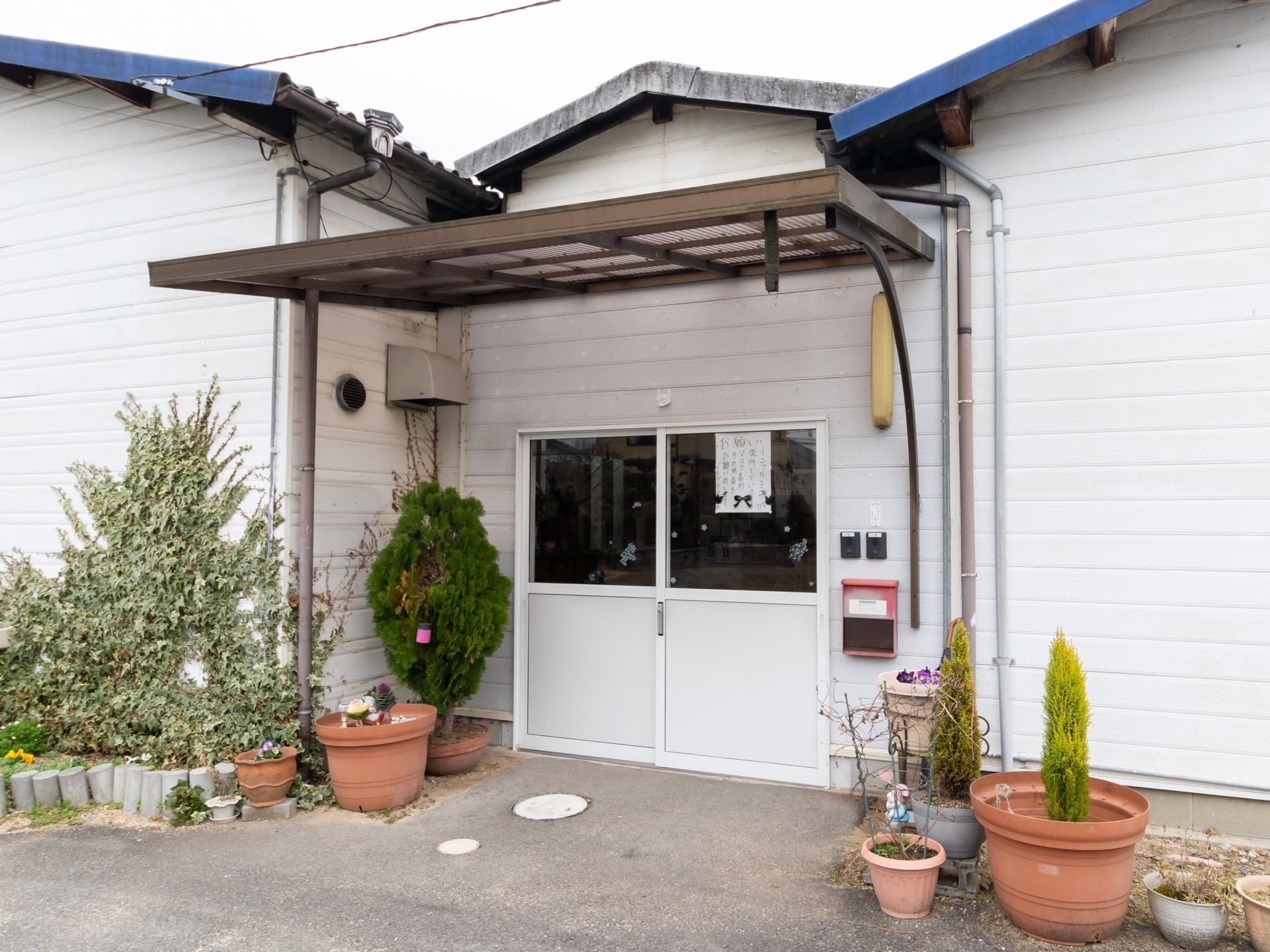 白い壁に、青や灰色の屋根の平屋の建物である。玄関のすぐ右側には、インターフォンや郵便受け、照明が付いている。