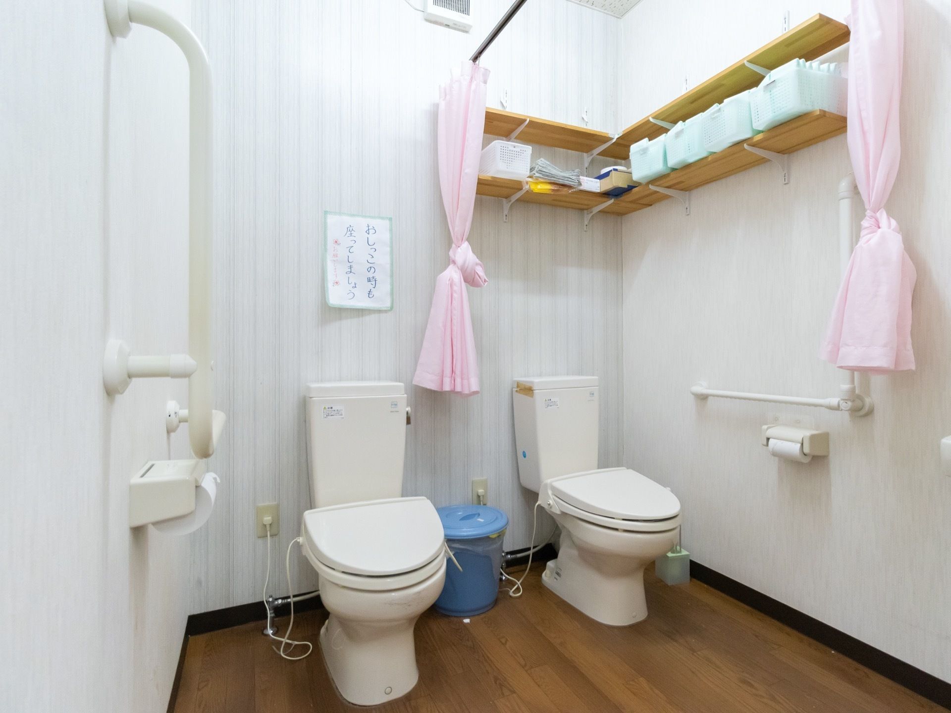 便座が2つ横並べに設置されており、その間にはブルーの蓋つきのゴミ箱が置いてある。天井には、カーテンが取り付けられている。