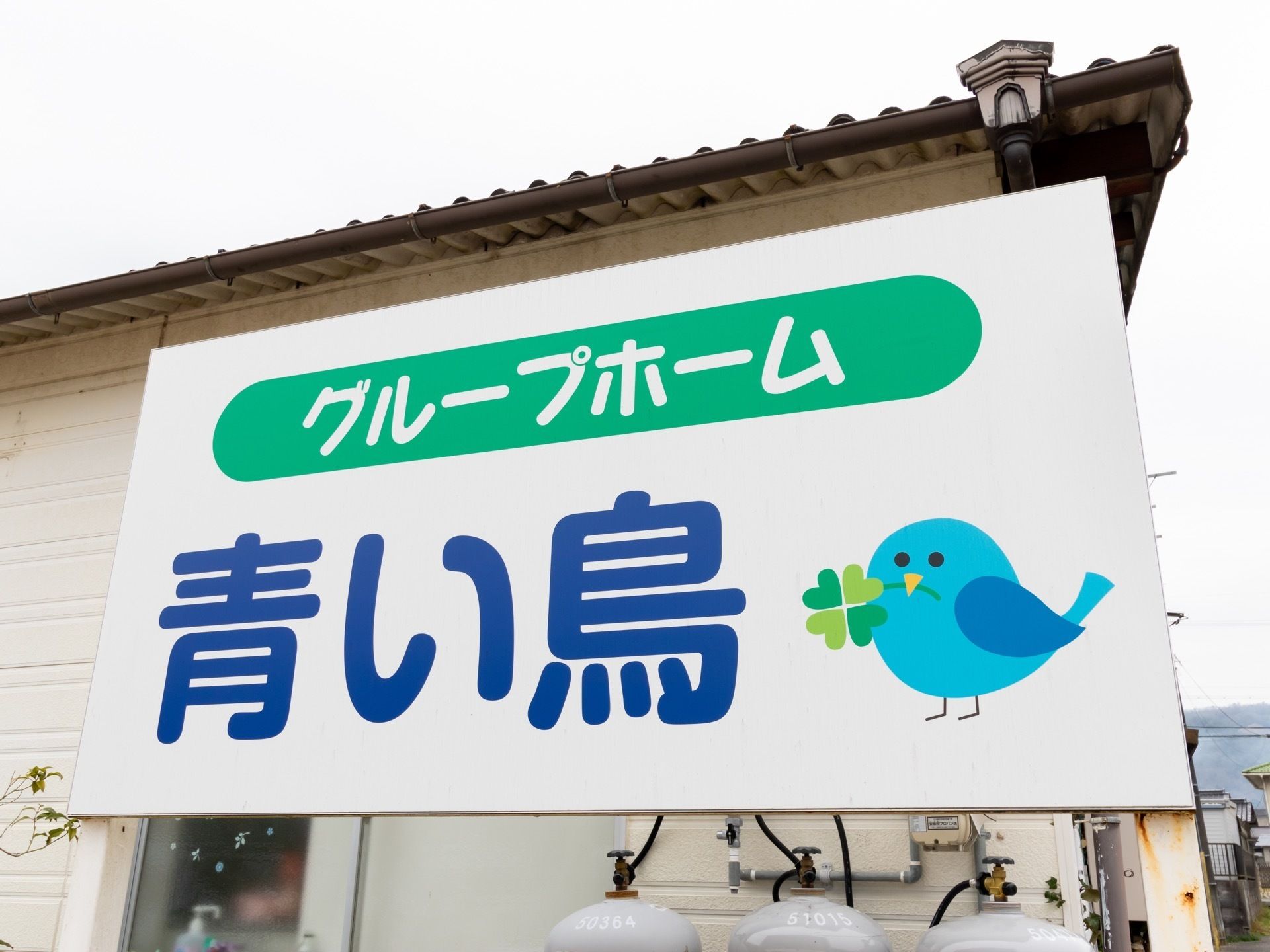 白い板に施設名や施設の種類が書かれており、クローバーをくわえた青い鳥の絵が一緒に描いてある。その下には、ボンベが3つある。