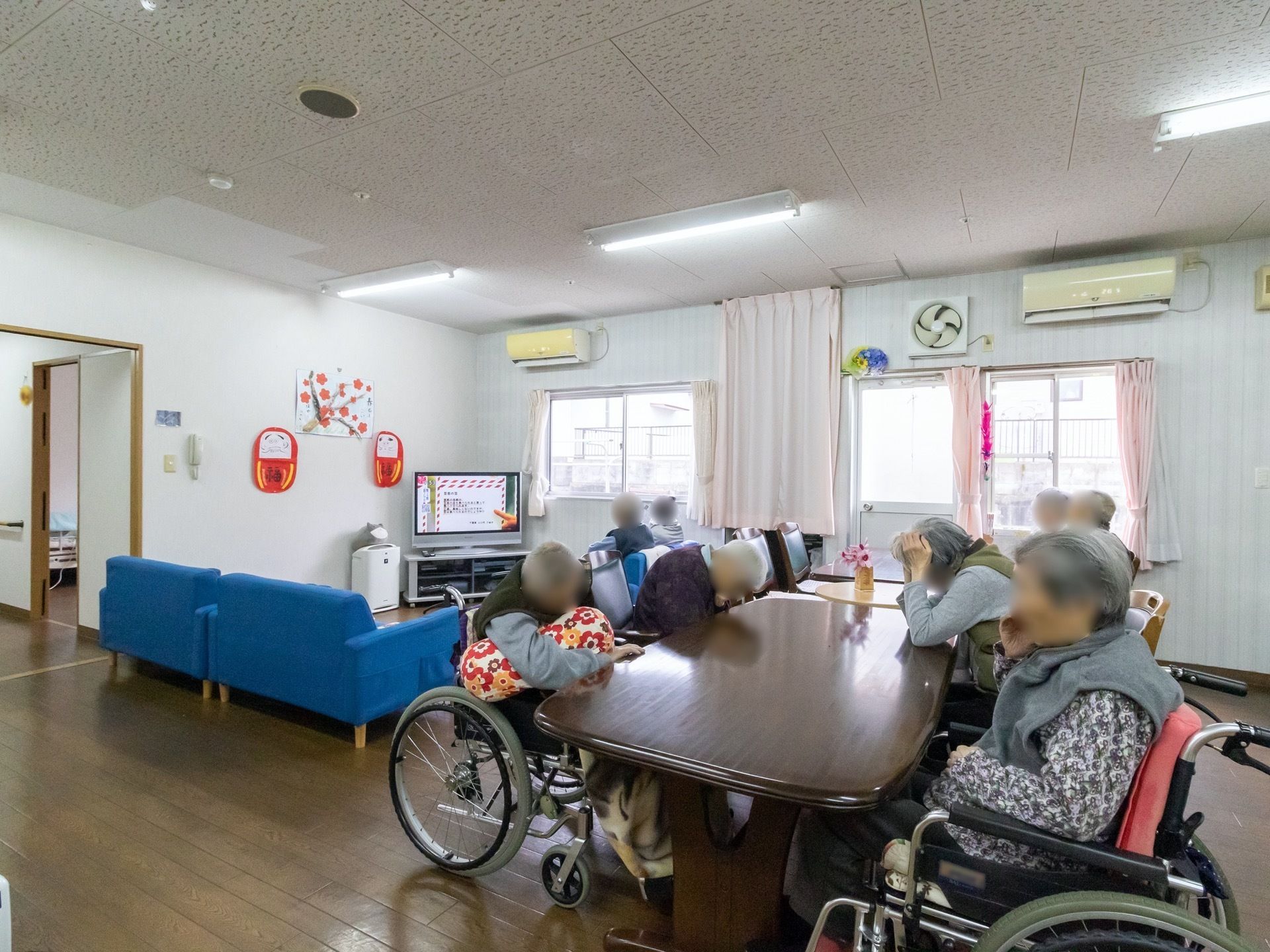 窓がある明るいリビング。左側にはテレビとともに青いソファが置いてある。右側にはテーブルが並べられ、複数の入居者様が椅子に腰かけている。