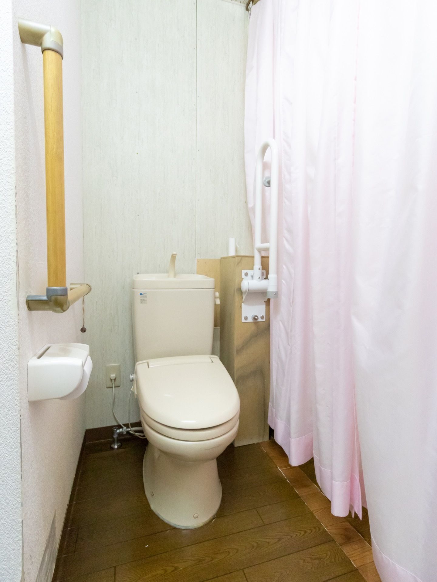 板張りの床でできた個室トイレである。左手の壁にL字の手すりが取り付けられ、右側はピンク色のカーテンが閉められている。