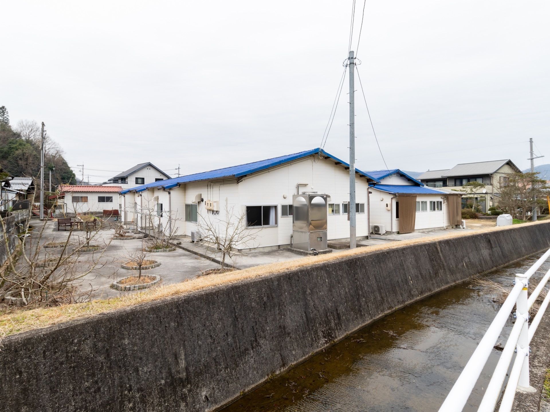 施設近隣の景色である。右奥に向かって小さな川が流れ、白いガードレールがある。皮の向こうには倉庫や住宅が並んでいる。