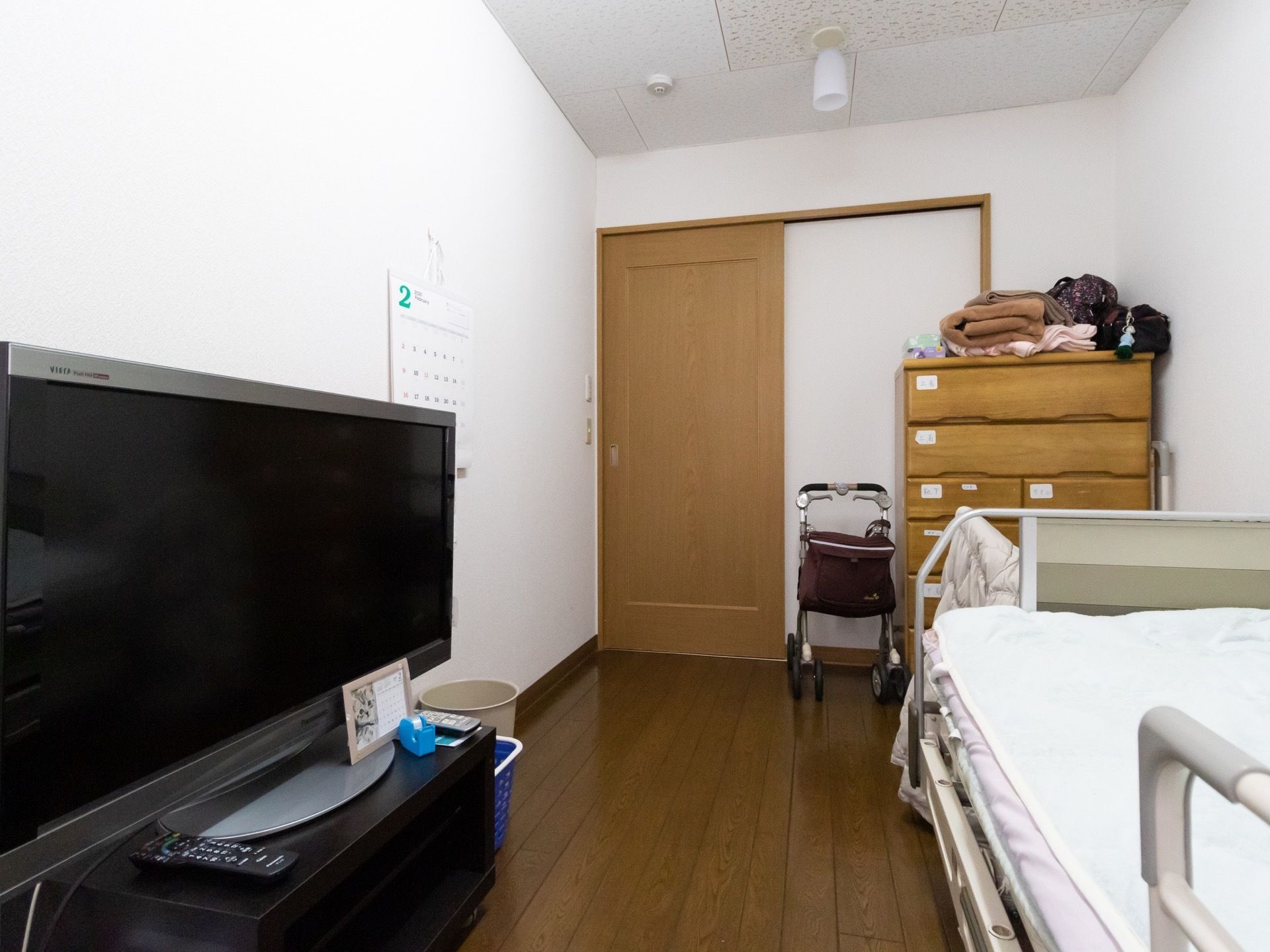 窓側から居室内を見た画像である。右側に介護用ベッドが置かれ、その前に大型液晶テレビがテレビボードにのせられている。