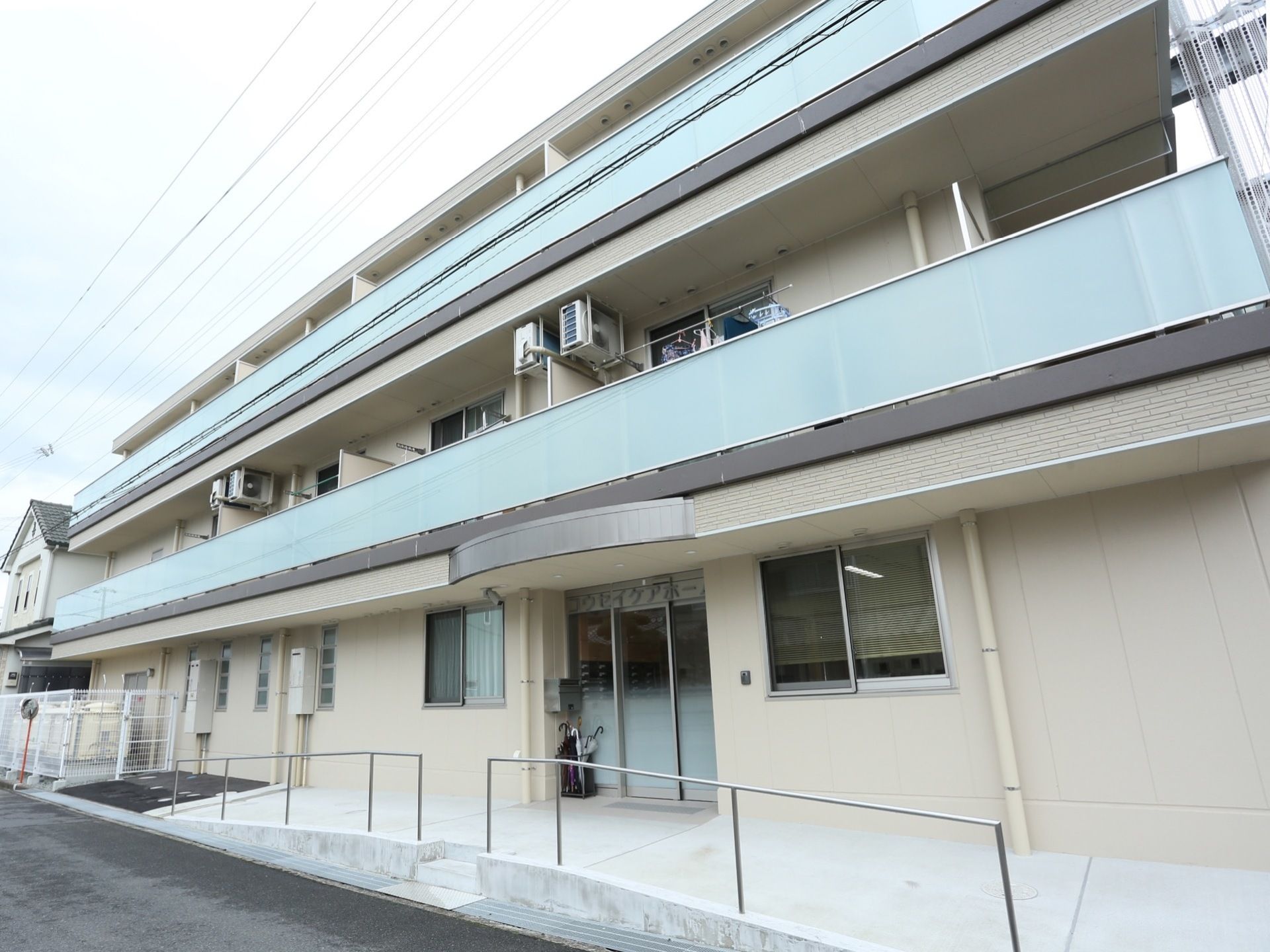 玄関前のアプローチには手すり付きのスロープがある。３階建ての建物で、２階以上にはベランダが設置されている。