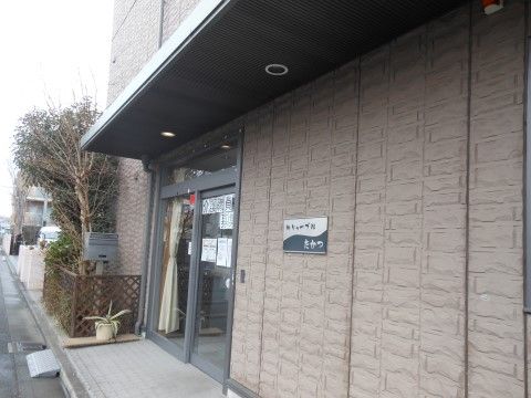 道路に面した茶系の3階建ての外観は落ち着いた雰囲気である。グレーの枠のガラス張りの玄関ドアの横側に、植栽の癒し空間を設けている。