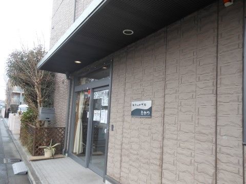 道路に面した茶系の3階建ての外観は落ち着いた雰囲気である。グレーの枠のガラス張りの玄関ドアの横側に、植栽の癒し空間を設けている。
