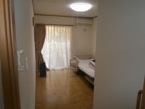 ワンルームタイプの居室である。床面はフローリングで段差がない。ドアは引き戸になっている。介護ベッド、エアコンが設置されている。