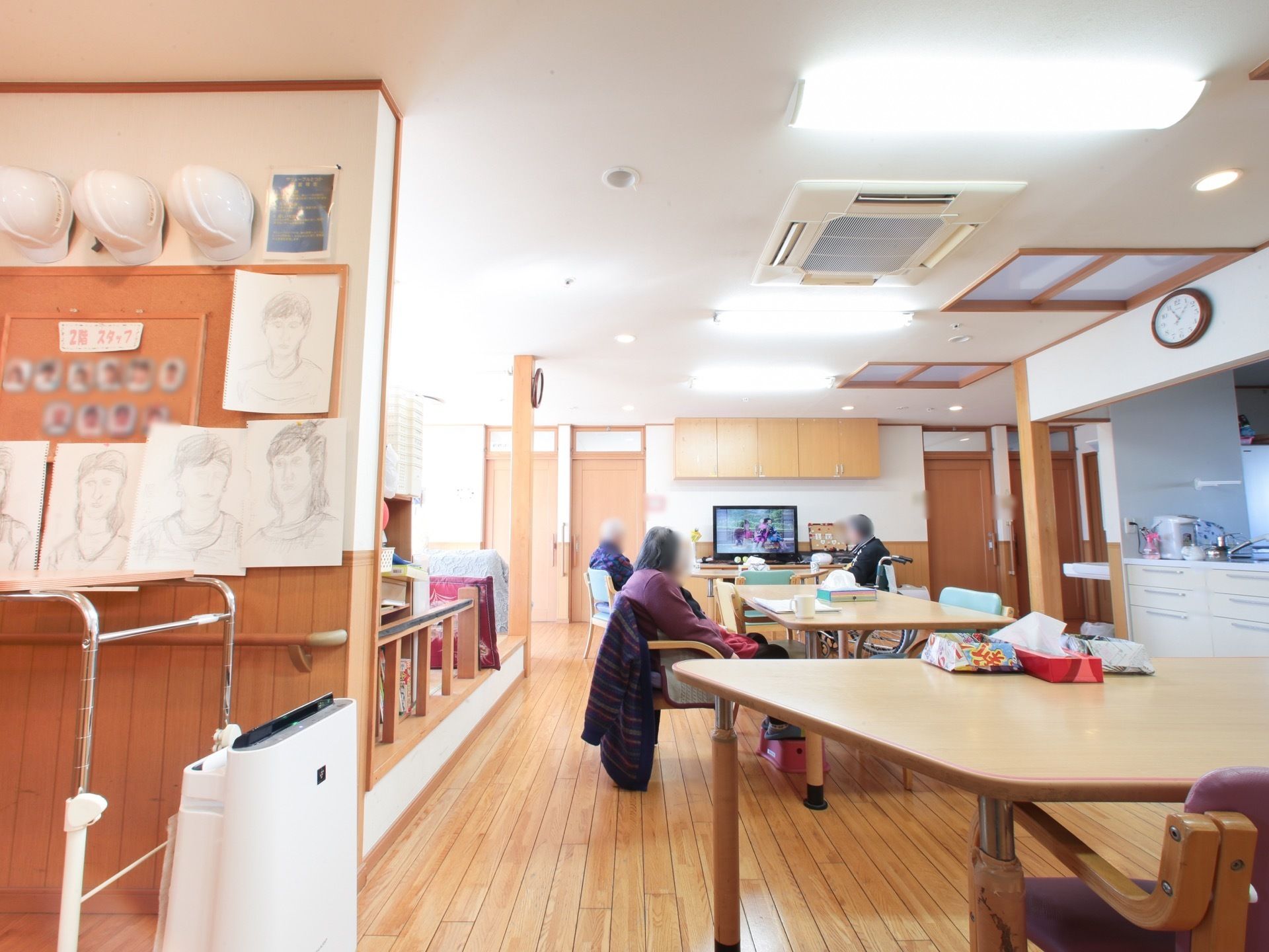 複数の入居者様が食堂に設置されたテレビを見ている。食事時間以外も食堂を利用できる。空気清浄機が設置されている。