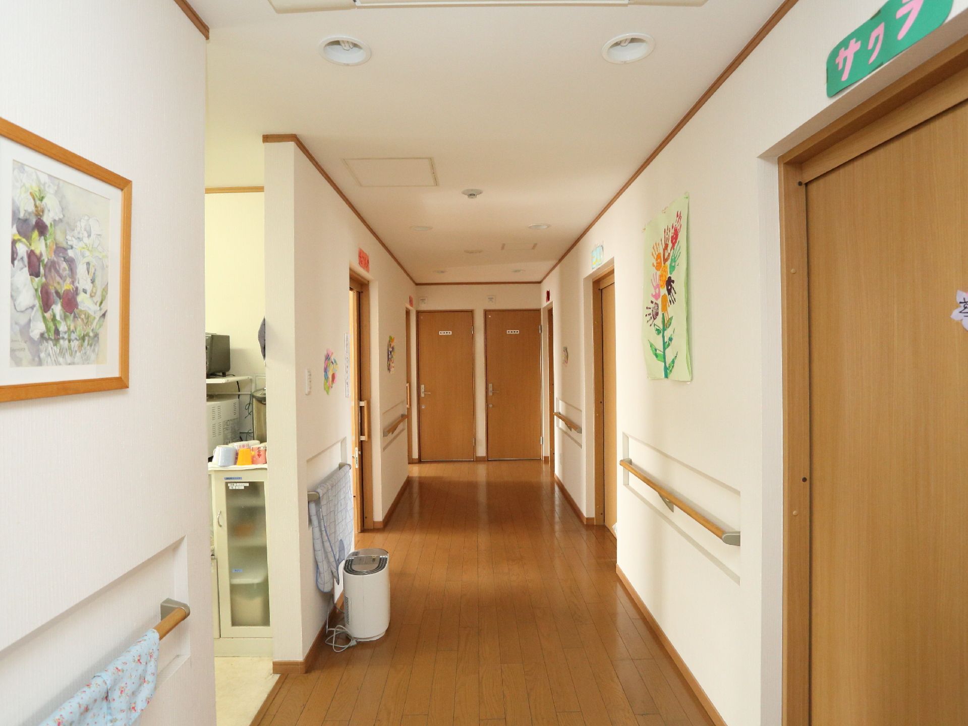 白い壁ときれいに磨かれたブラウンの床が明るく清潔感のある廊下。壁には花の絵やペーパークラフトが飾られている。
