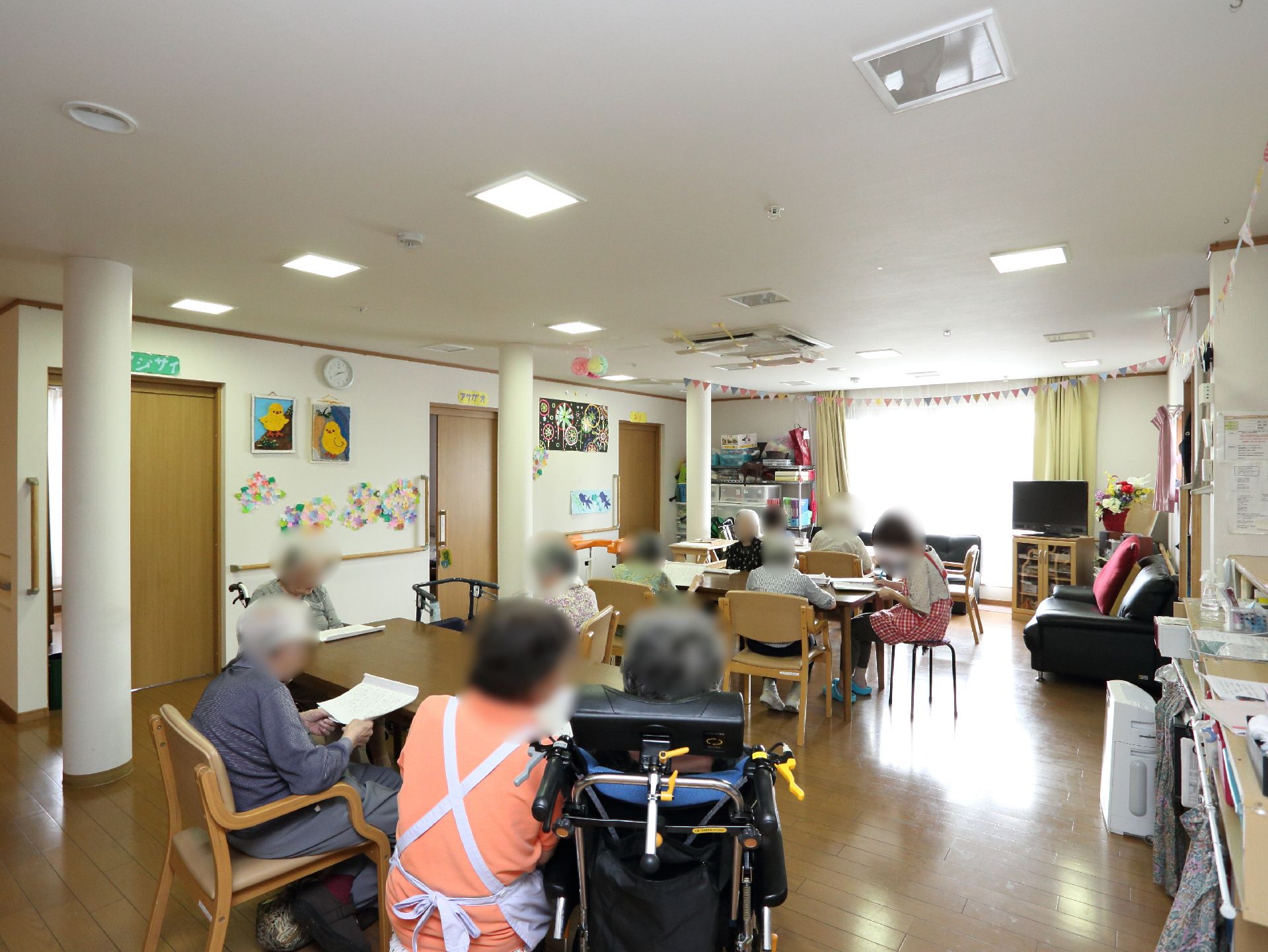 開放的な共有フロアには入居者様とスタッフが集まっている。テーブル、椅子、ソファが置かれ、リラックスできる雰囲気。