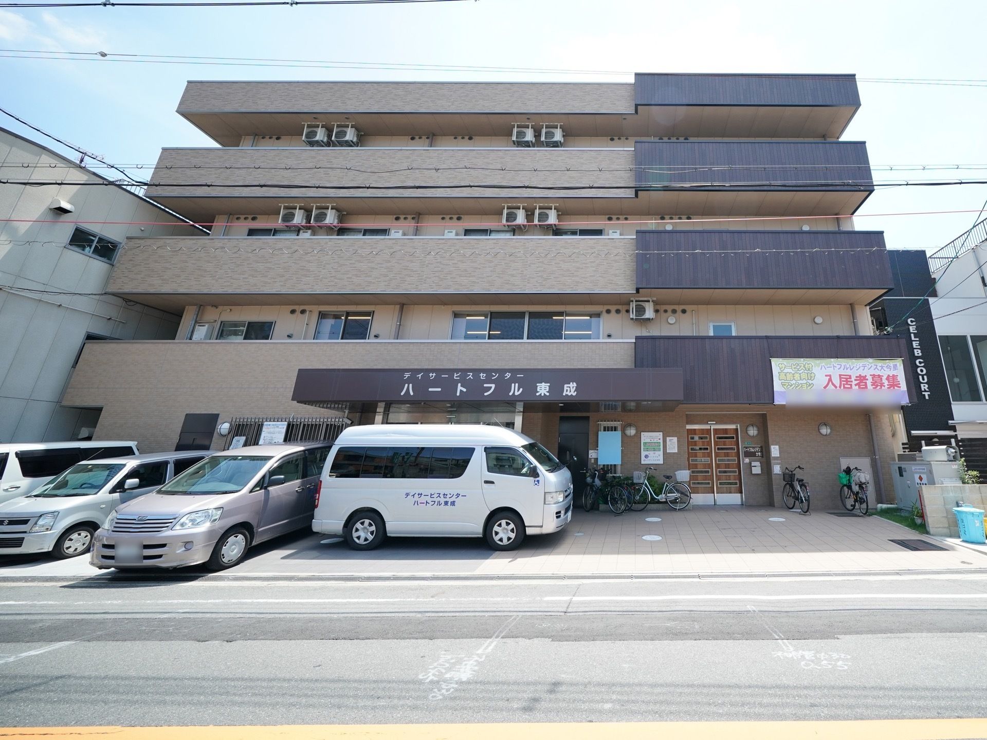 建物の前が駐車スペースになっており、車が数台停められている。２階のベランダに入居者様募集の垂れ幕が掛けられている。