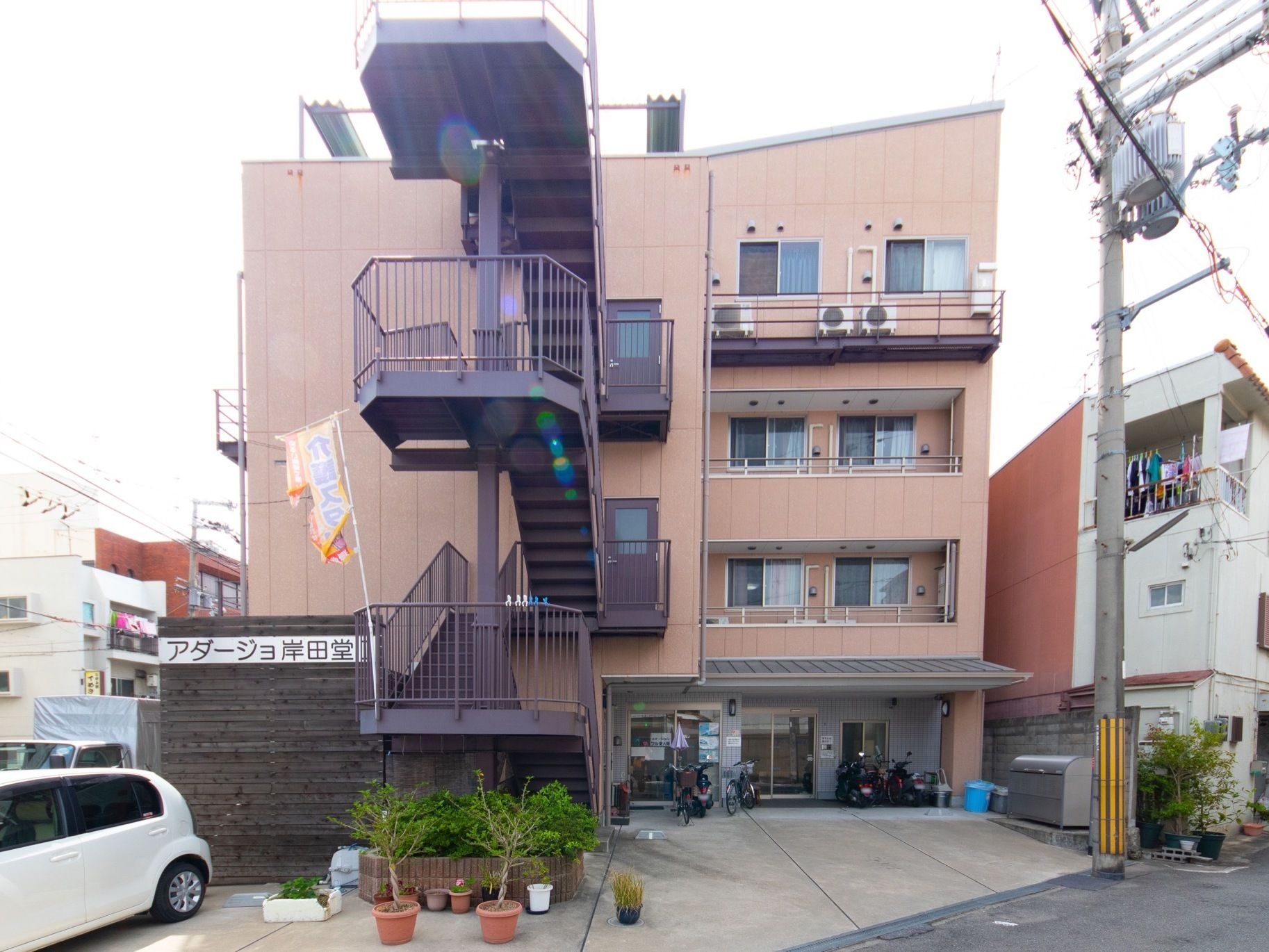 薄いブラウンを基調とした４階建ての建物である。横側に非常階段を設置している。その下に低木を植えた鉢とプランターを置いている。