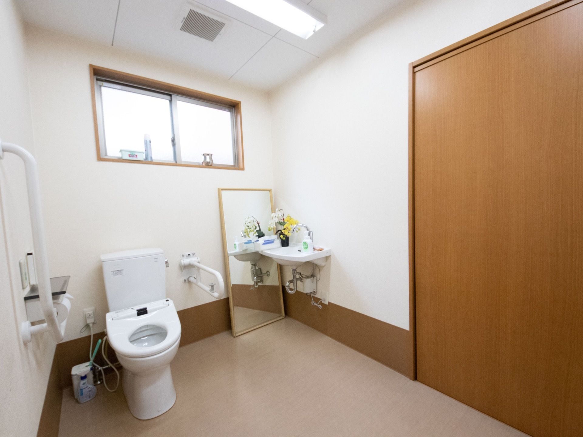 洗面台を備えた広々としたトイレスペースである。上部に窓があり、下に大きな鏡を置いている。便座の両側に手すりを設置している。