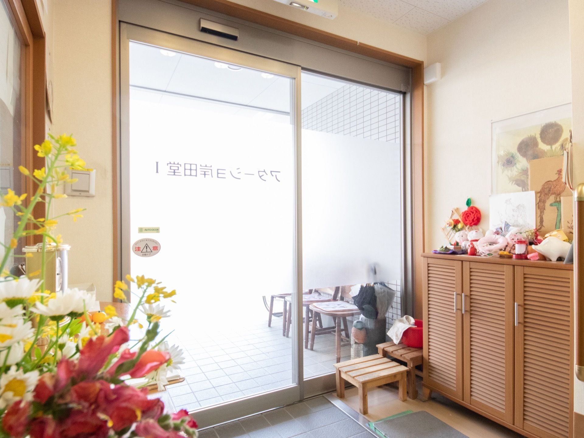 玄関の出入口の自動ドアに施設名を表示している。入って左側に木目調の収納庫を備え、上に置物などを飾っている。