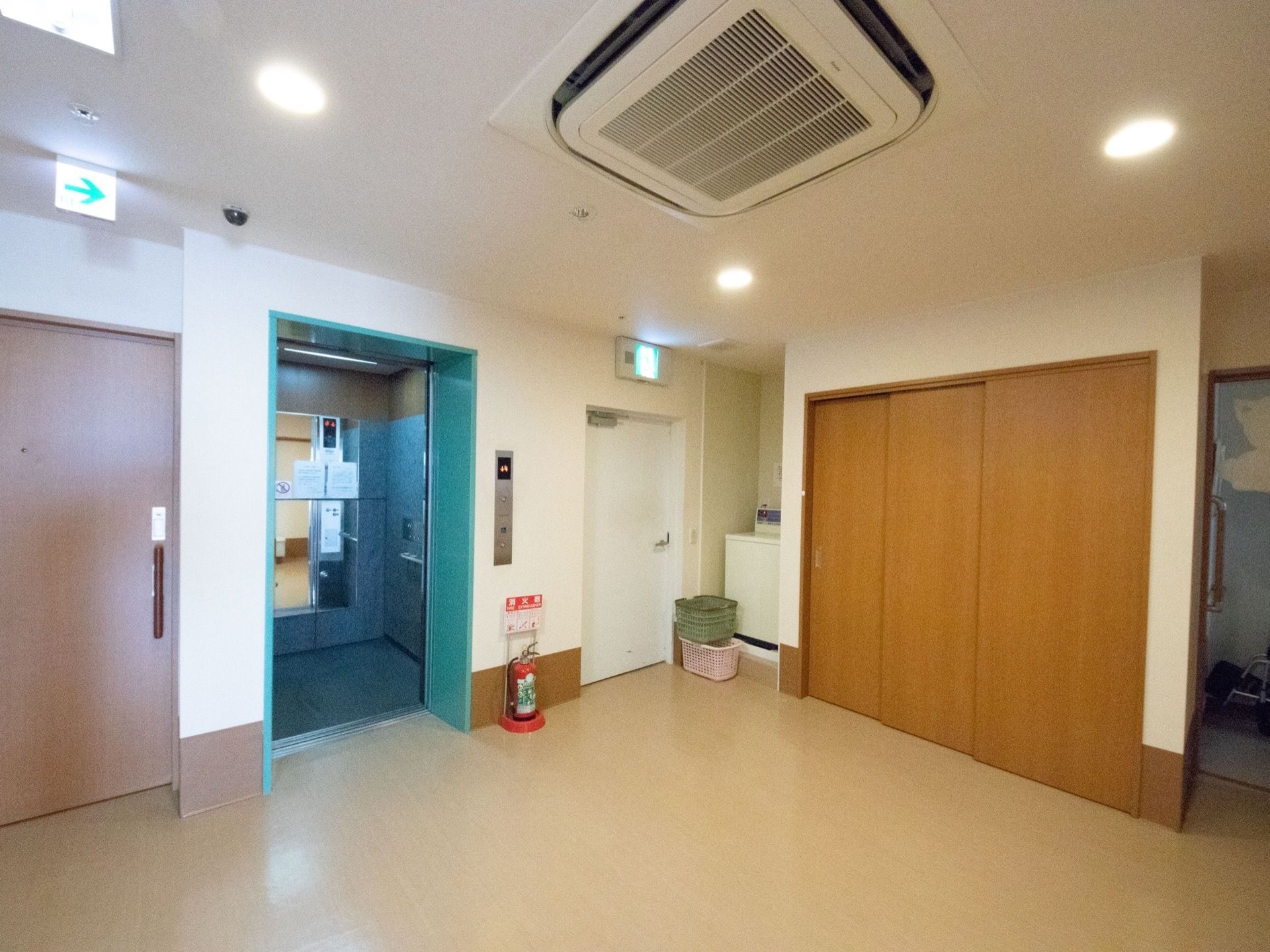 広々としたエレベーターホールである。天井には、大きめのエアコンを備え、ドアの近くに消火器を設置している。