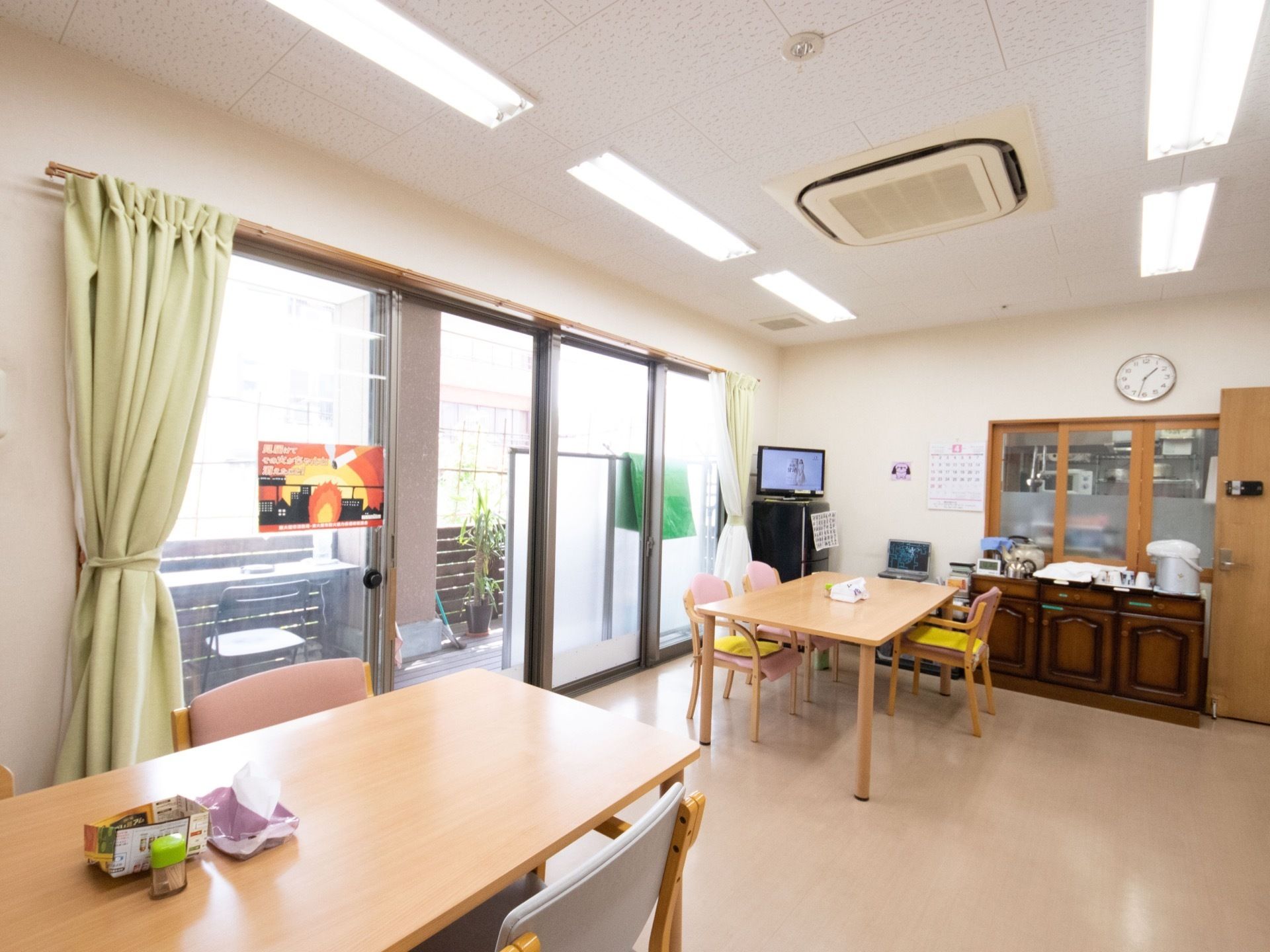 共有スペースの角にテレビを備え、横の収納棚の上にポットや湯飲みなどを置いている。壁にカレンダーや時計を掛けている。