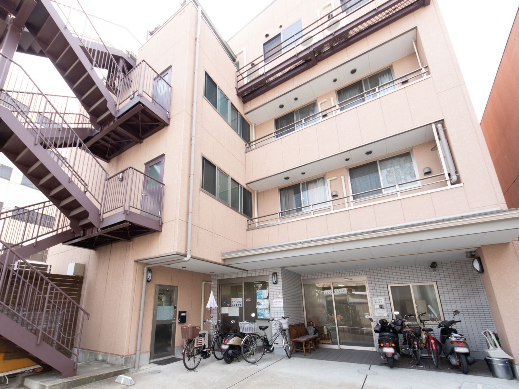 薄い茶系のL字型構造の建物である。玄関横の壁際に椅子と傘立てを置いている。バイクや自転車が何台か停まっている。
