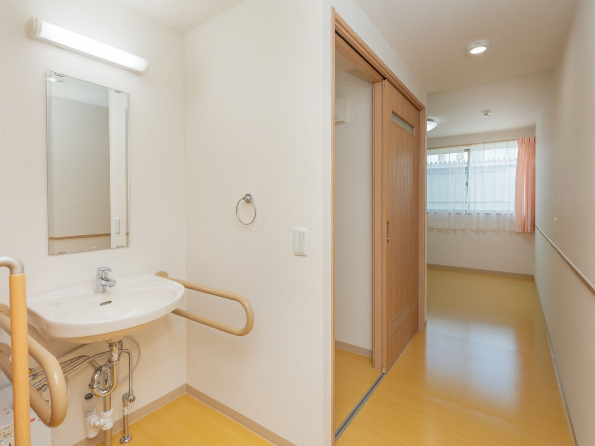 居室はフローリングである。縦長の鏡、照明が設置された洗面台があり、両側には手すりが取りつけられている。