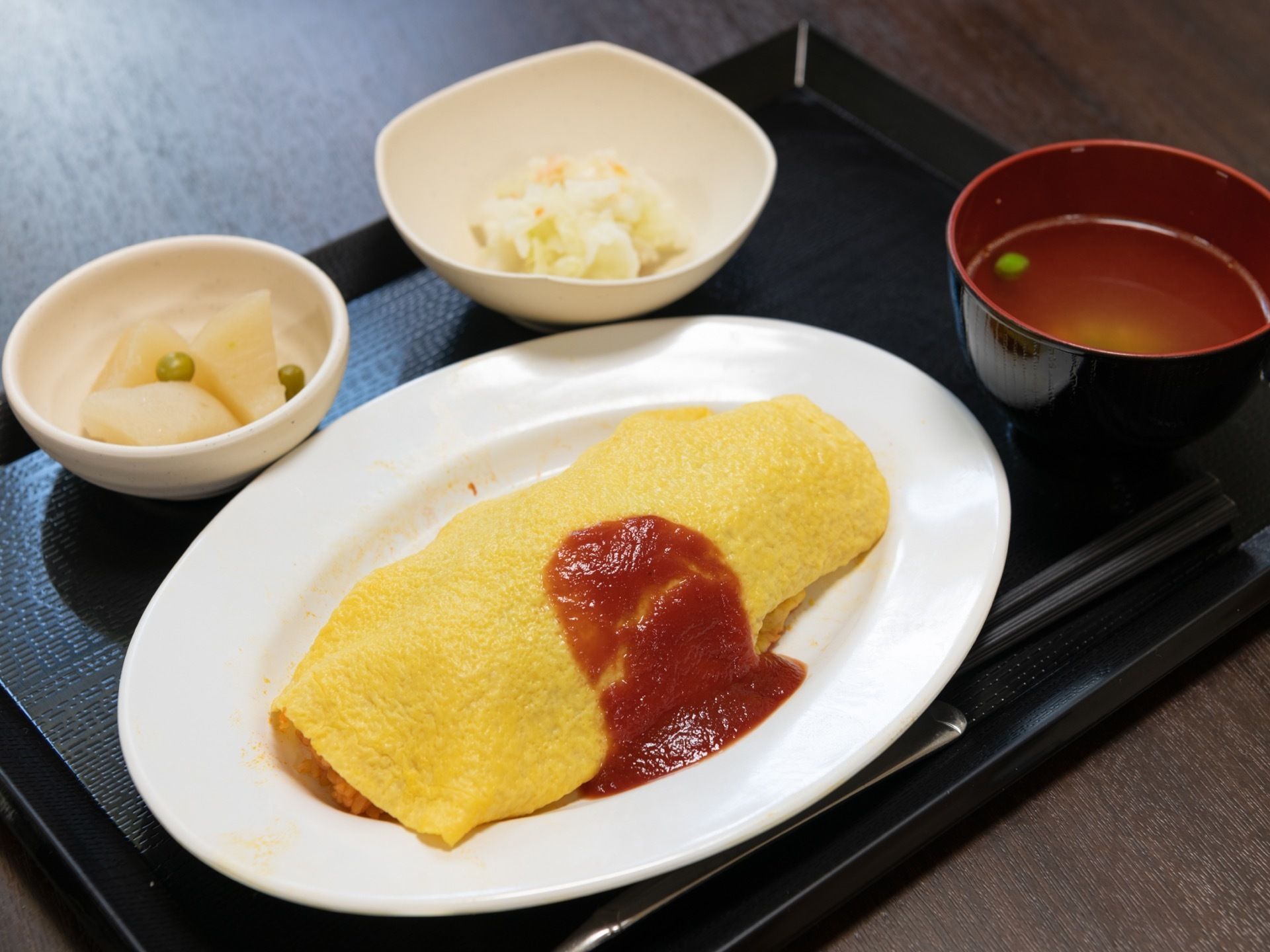 黒いお盆の上に食事が配膳されている。メインはオムライスである。他にお味噌汁と2品のおかずが用意されている。