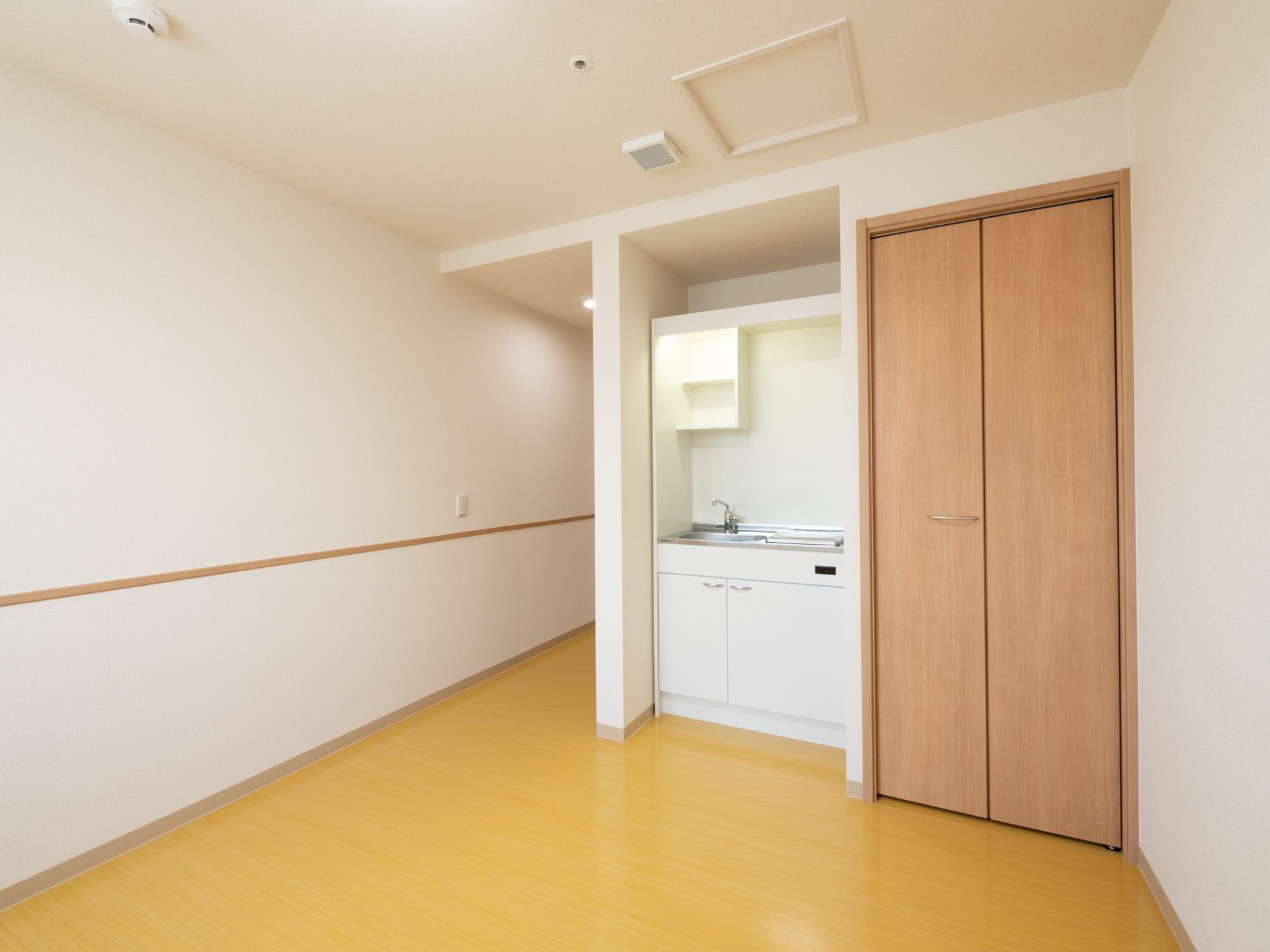 居室にはナチュラル色のフローリングが敷かれている。白いキッチンスペースがあり、その横には扉付きの収納スペースが設置されている。